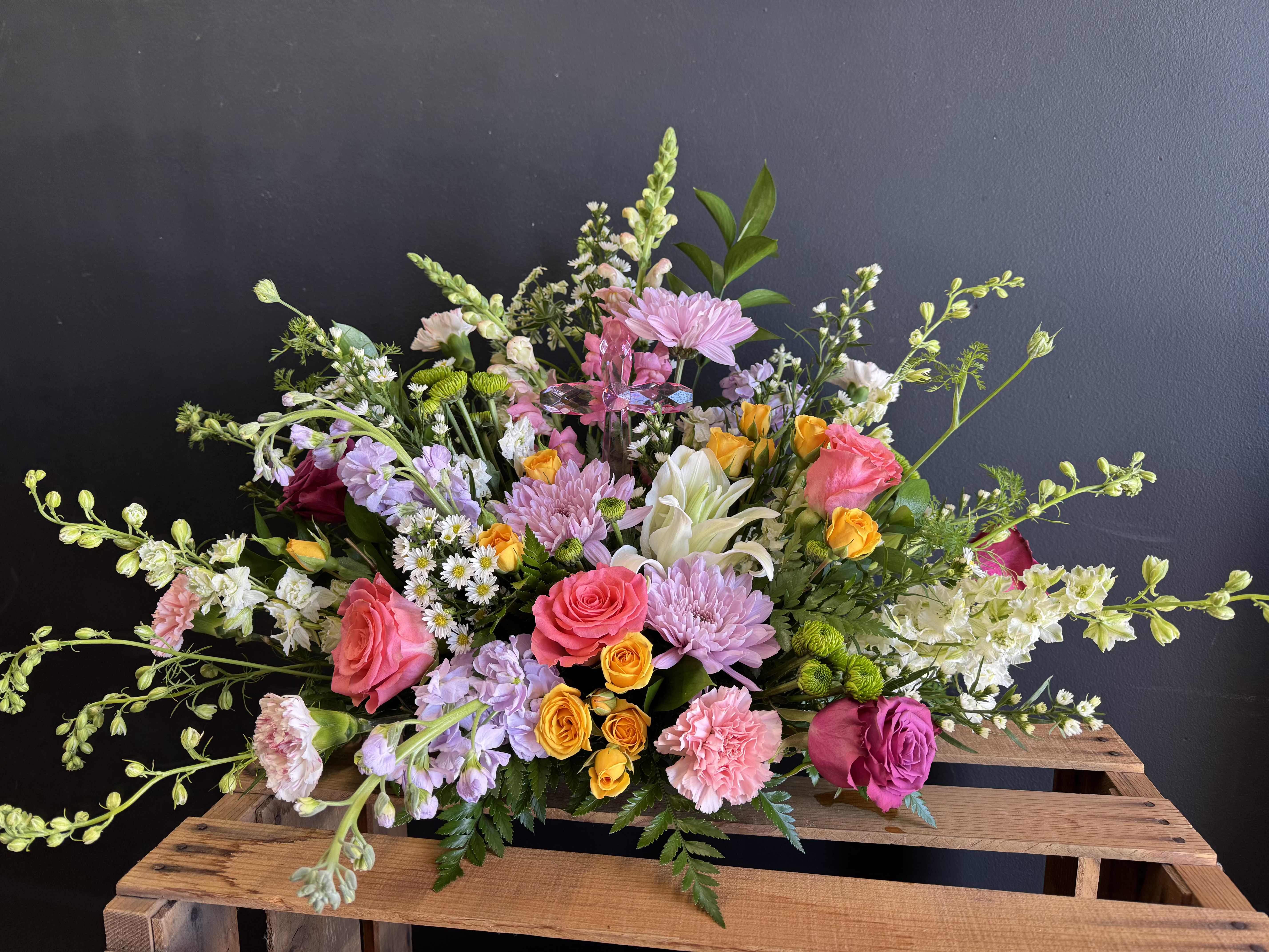 Springtime Serenity - A lush garden-style arrangement featuring pink roses, lilies, snapdragons, carnations, and whimsical seasonal blooms in soft pastel shades. Designed with airy texture and fresh spring color, this arrangement brings beauty and comfort to any occasion. A small cross is added in the middle for a forever keepsake. 