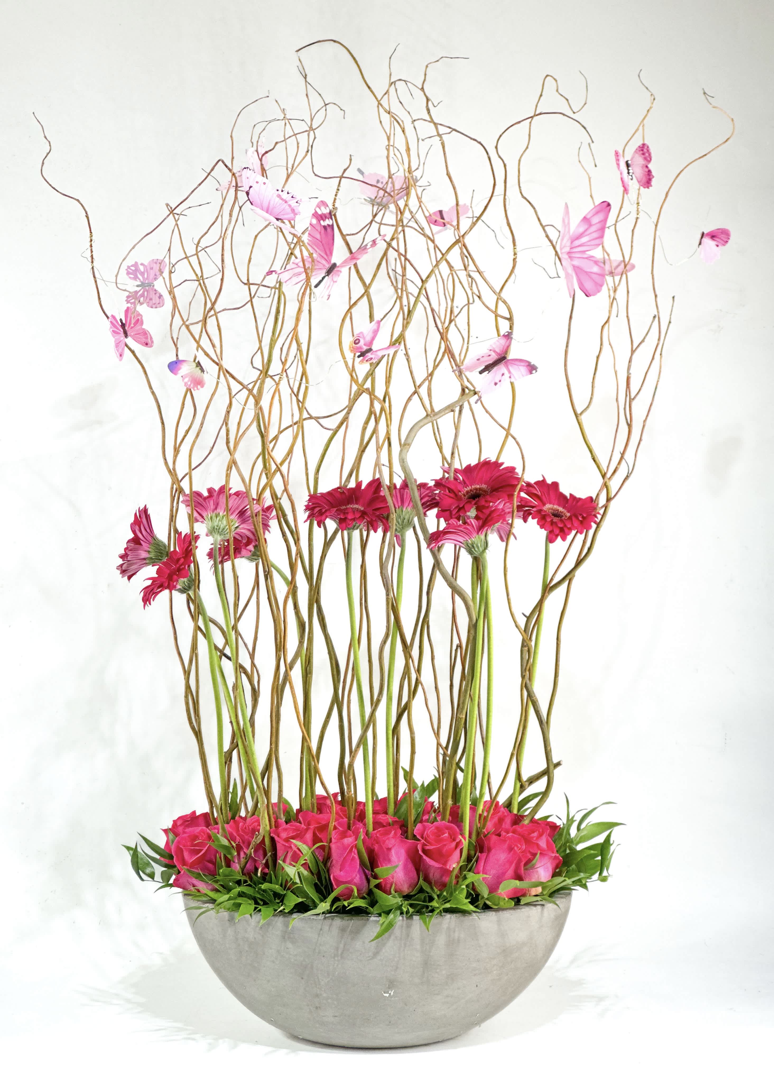 Pink roses and gerbera daisies in a gray bowl with tall curly branches and butterflies
