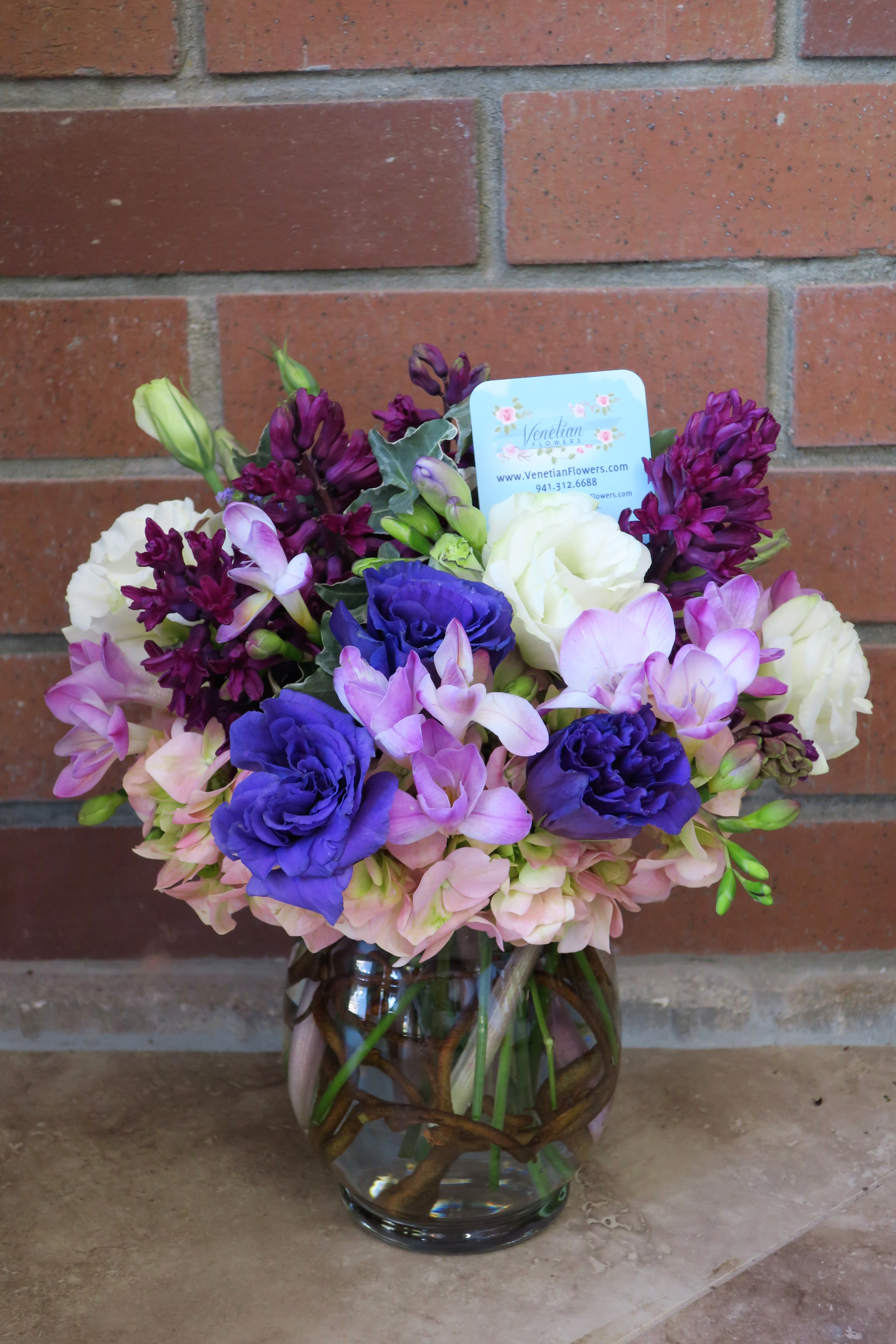 Round vase arrangement with purple, pink, and white flowers in a clear vase with branches inside