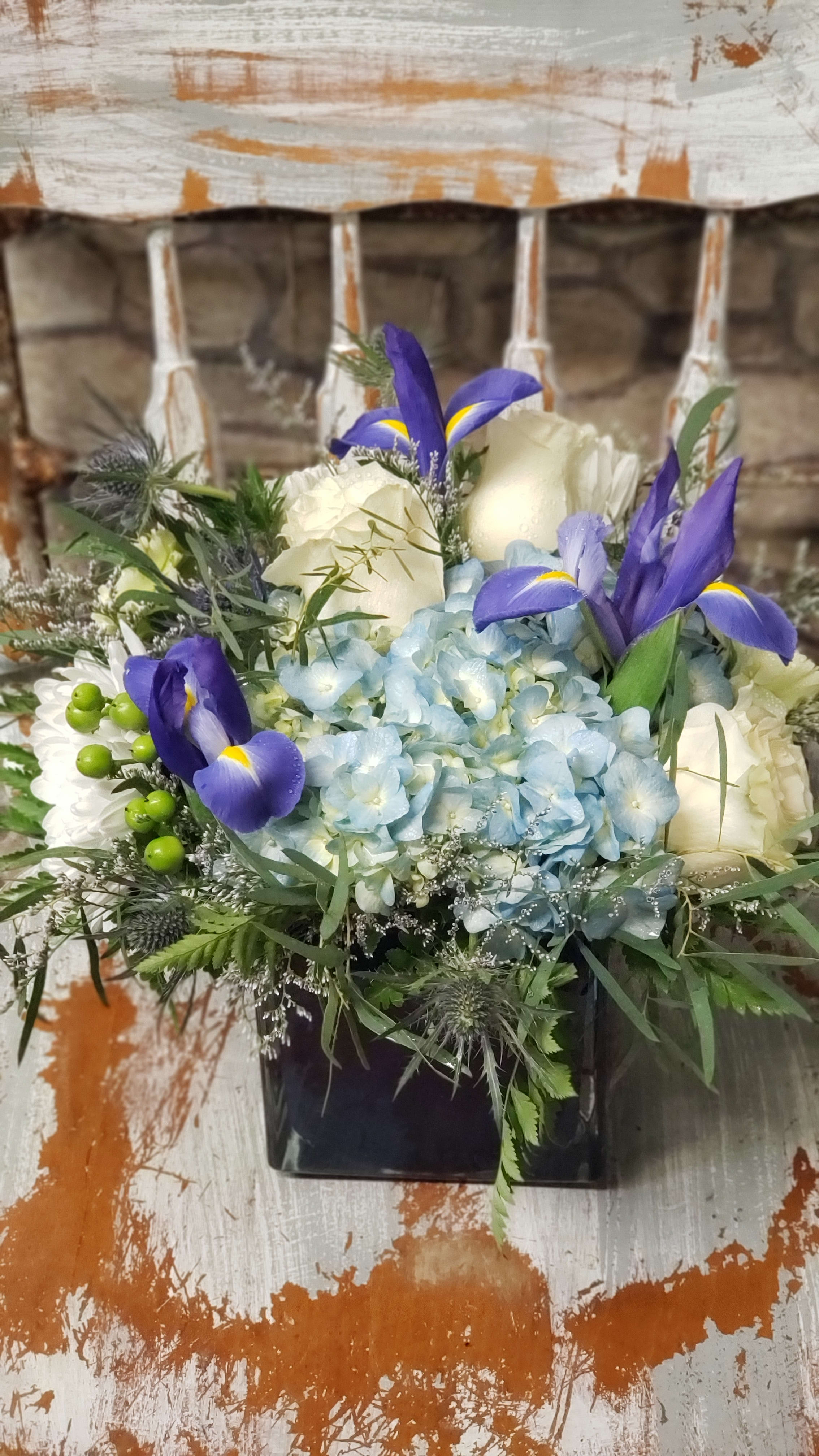 Compact arrangement of blue hydrangea, white roses, and purple iris in a dark cube vase