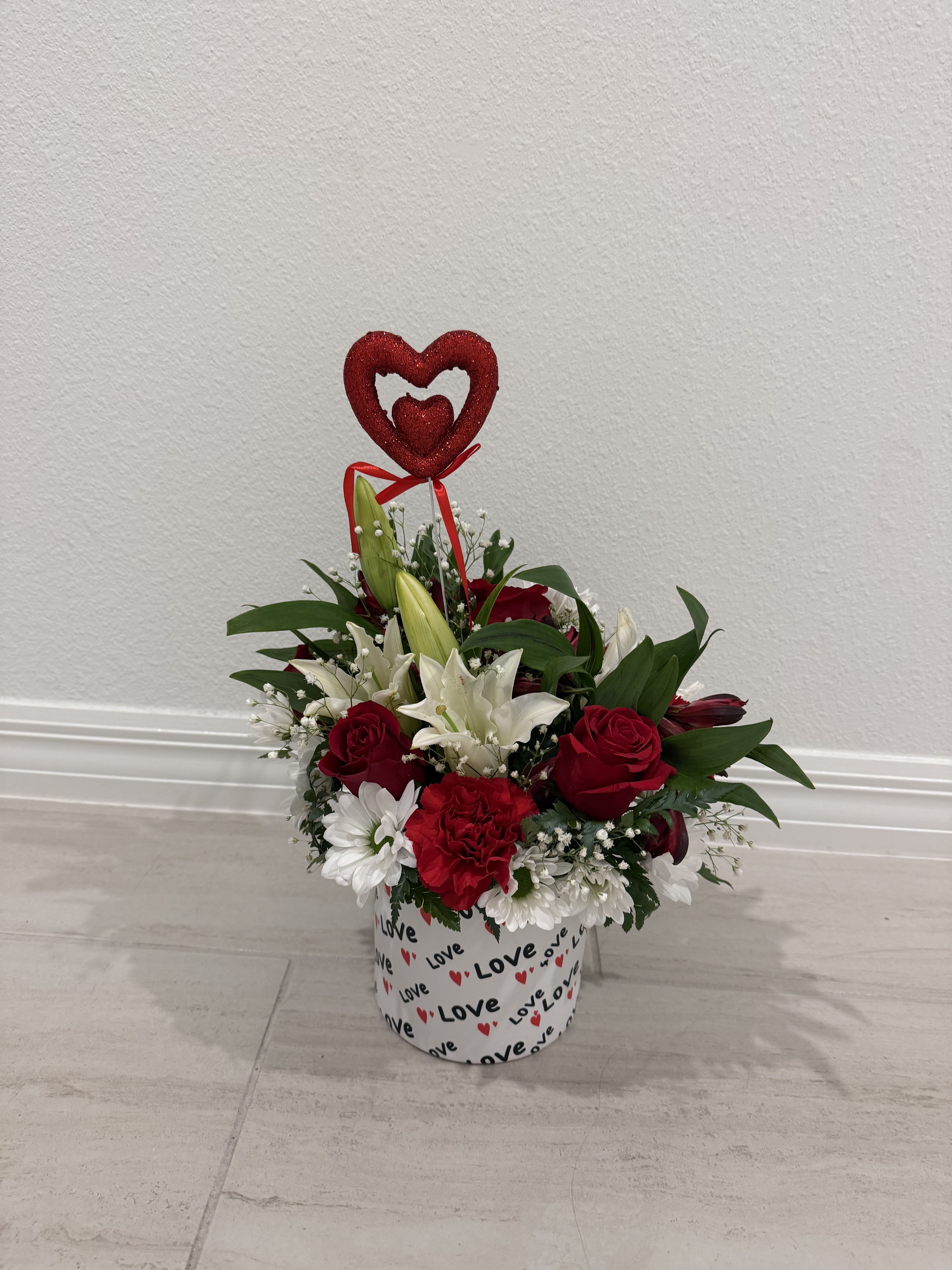 Red and white floral arrangement in a heart-decorated box with a heart topper