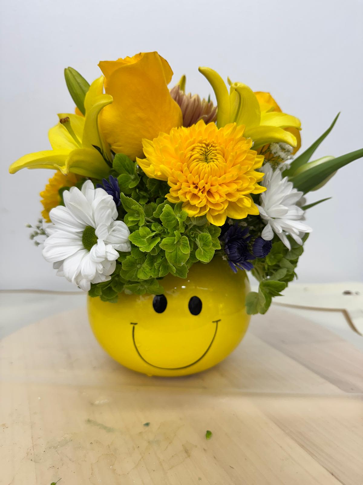 Yellow smiley-face container holding a mixed bouquet of yellow, white, and purple flowers