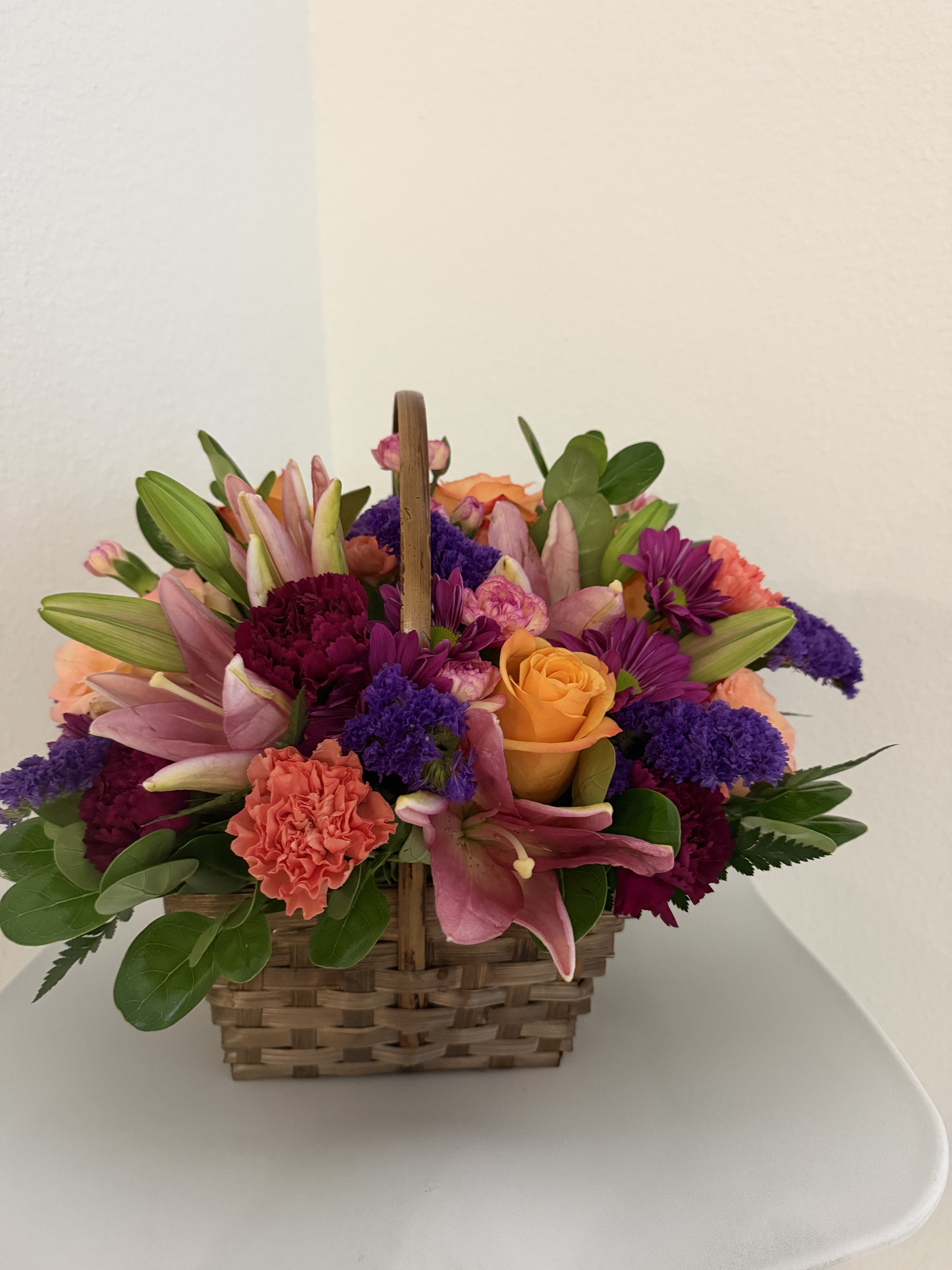 Golden Bloom Basket - A radiant mix of peach lilies, vibrant orange roses, purple fillers, and soft pink carnations arranged in a charming woven basket with fresh greenery. A bright and joyful design perfect for celebrating any special moment.