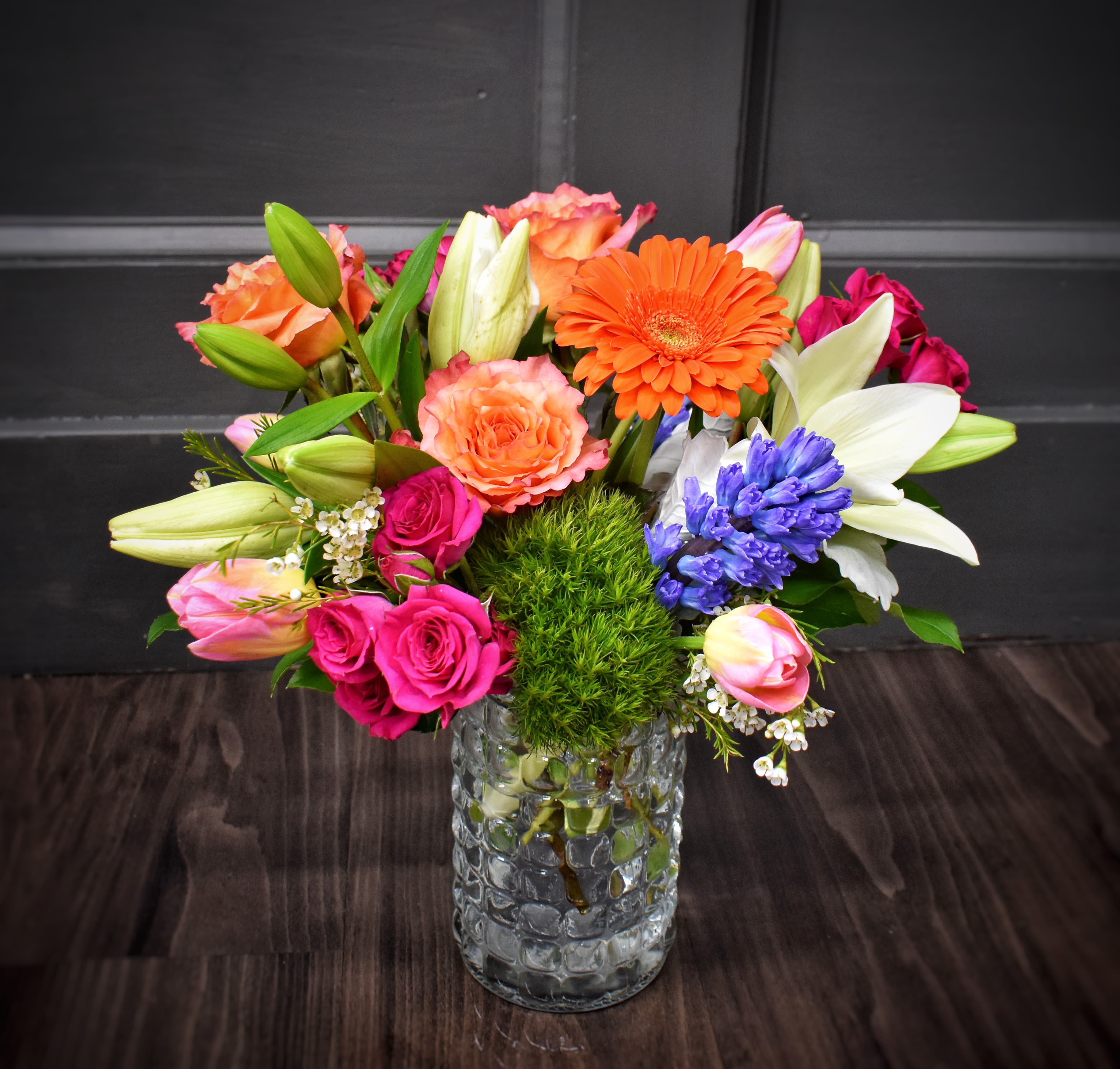 Symphony of Spring- PREMIUM - A burst of springtime cheer-bright pink roses paired with zesty orange gerbera daisies and soft white lilies create an uplifting birthday bouquet or just-because arrangement that makes the person you're thinking of feel genuinely celebrated. This flower arrangement for birthdays, congratulations, or everyday surprises balances warm and cool tones-pink, orange, white, and purple-for a lively, polished look that reads joyful without being over-the-top.  Built as a rounded, full composition, it features purple hyacinth and pink-and-cream tulips woven among the roses, gerberas, and lilies to add texture, scent, and seasonal charm. Arranged in a clear textured glass vase, the display is ready-to-gift and looks great on a dining table or office desk; the vase keeps stems neatly supported and showcases the colorful stems. This is the kind of bouquet that says "you matter" in a bright, confident voice-perfect for friends, coworkers, or family when you want them to smile.