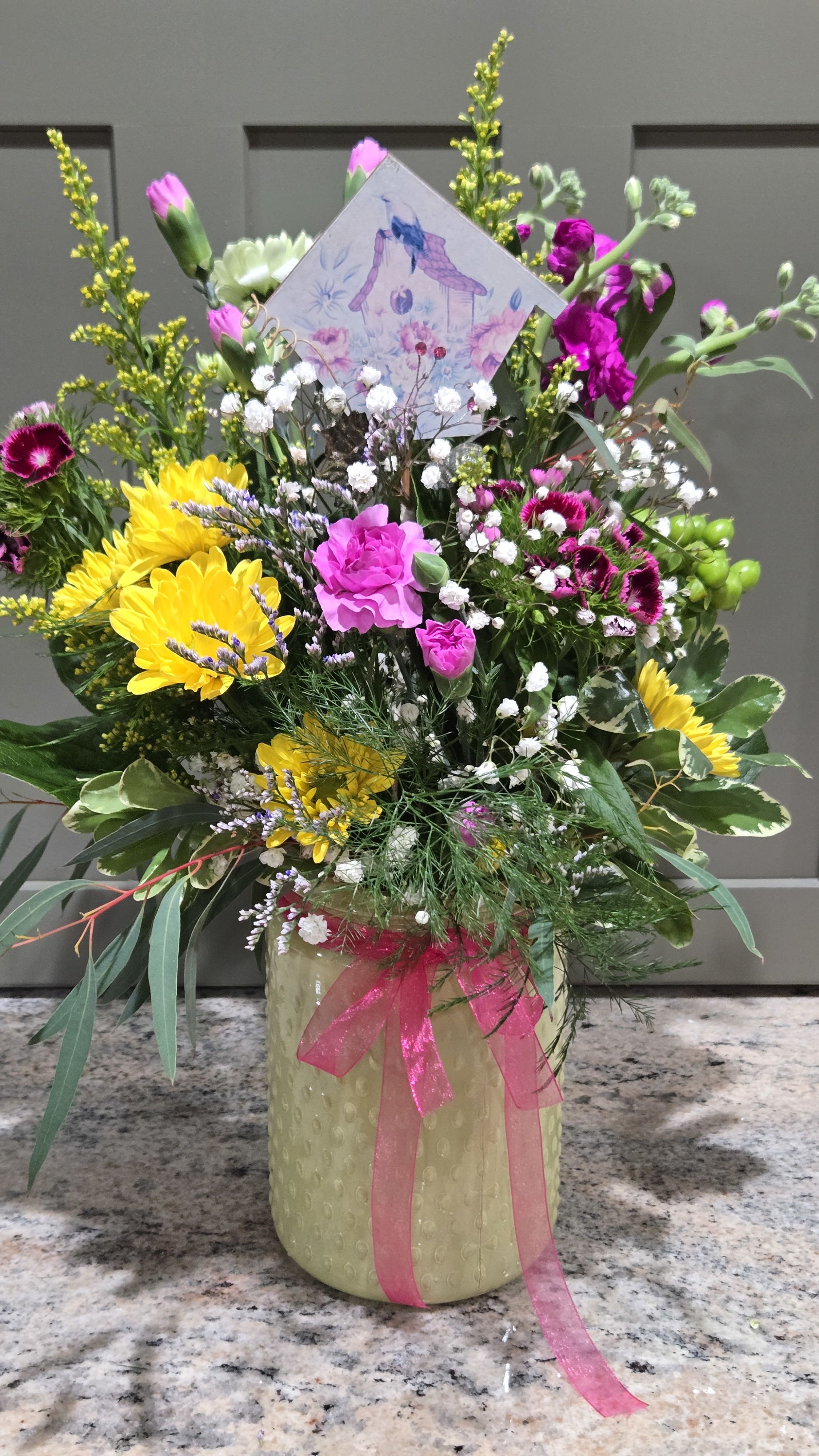 Sunny Blooms - Sunshine-bright yellow chrysanthemums and hot pink carnations give this mixed flower arrangement an instant lift-perfect as a Mother's Day bouquet, birthday surprise, or just-because pick-me-up for someone who loves cheerful color. The tall, abundant design arrives in a pale yellow glass vase topped with a sheer pink ribbon and a tucked greeting card, so it reads like a ready-to-give gift that will make her feel truly seen. The vase is textured and tied with a coordinating pink ribbon; a decorative pick is included. Its lively palette suit springtime celebrations, housewarmings, new baby visits, retirements, thank-you gestures, get-well wishes or simply saying "thinking of you." Care: keep flowers in fresh water, trim stems and refresh water every few days to extend the joyful display.
