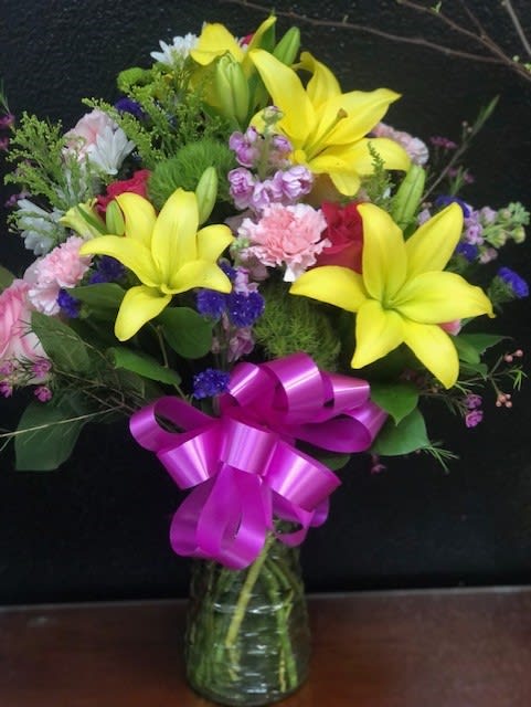 Bouquet of yellow lilies, pink flowers, and a purple ribbon in a glass vase