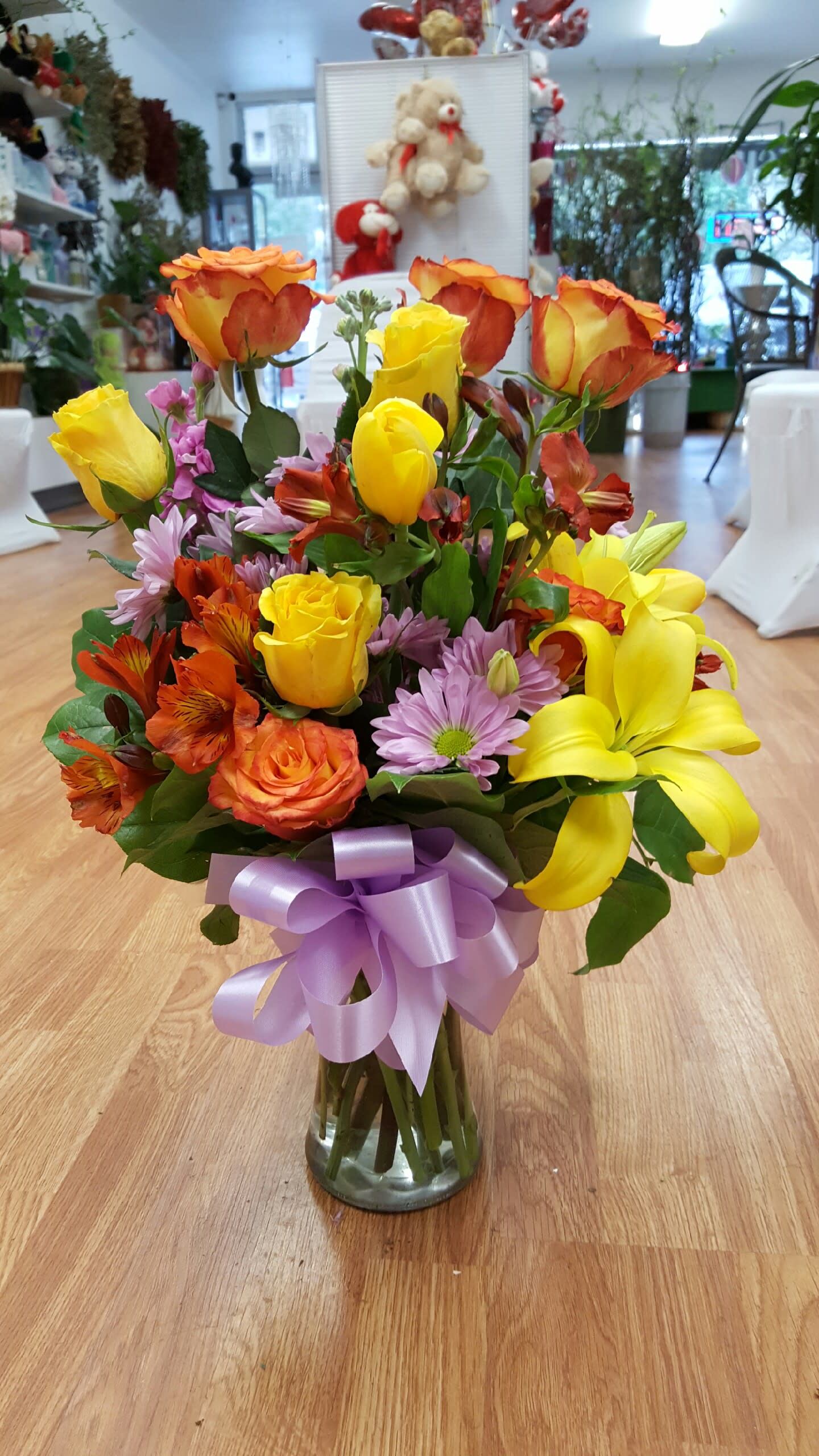 Mixed bouquet of yellow and orange roses with lilies and purple daisies in a glass vase