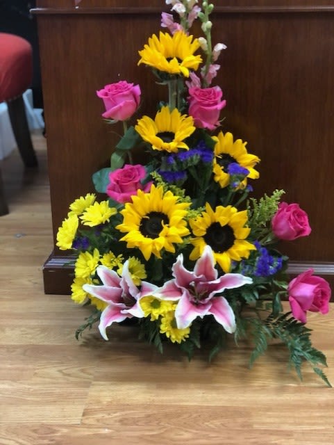 Bright and Snazzy Design - Sunflowers, Roses, Daisies, Static, Snap Dragon, and Stargazer Lilies with mixed greens arranged in floral foam to create a beautiful front-facing design
