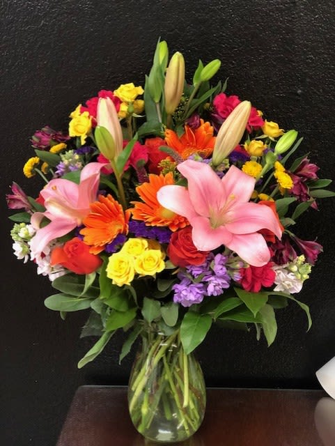 Blossoming Briliance - A vibrant floral arrangement designed to bring joy and color to any occasion. This bouquet features a stunning mix of bold and cheerful blooms, including large pink lilies, bright orange gerbera daisies, and hot pink and yellow roses. Clusters of purple and white stock flowers add rich texture, while assorted greenery provides a lush base. The entire arrangement is presented in a classic clear glass vase, making it a perfect centerpiece for a celebration or a thoughtful gift to brighten someone's day.