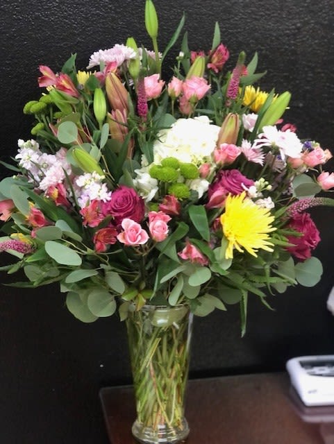 Garden Of Joy - This vibrant bouquet features a lush mix of flowers, including a striking yellow spider mum, deep fuchsia roses, and soft pink alstroemeria. White hydrangea and clusters of purple and white stock add texture and depth, while unopened lily buds promise future blooms. The arrangement is artfully presented with eucalyptus greenery in a clear glass vase, creating a beautiful and cheerful centerpiece.