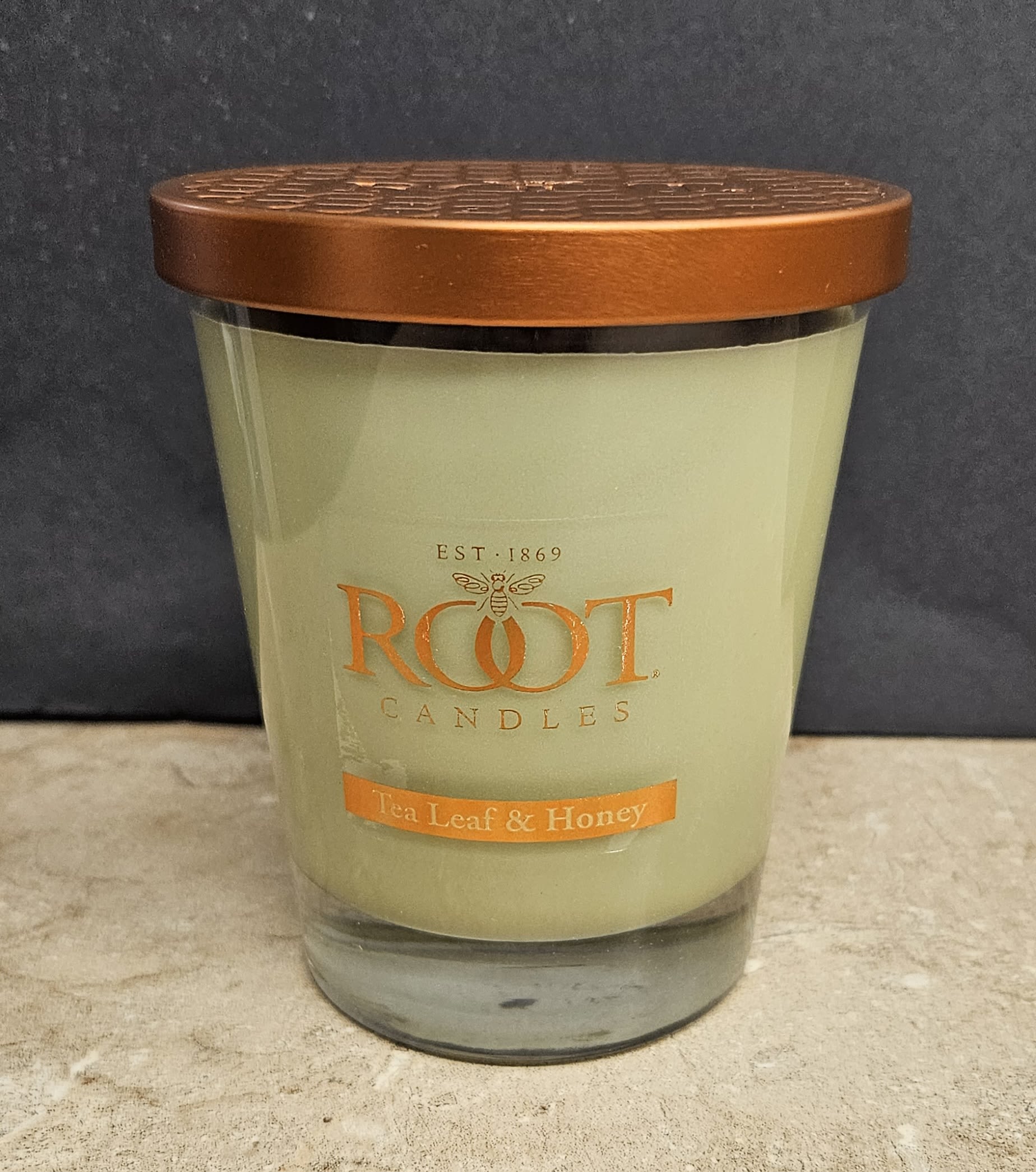 Root Candle Tea Leaf &amp; Honey M - Warm, honeyed notes and a soft, creamy glow make this scented candle a thoughtful gift for someone who cherishes cozy home moments - ideal as a housewarming present, birthday surprise, or a simple "thinking of you" gesture. As a home fragrance gift rather than a floral arrangement, it brings the calm of tea and honey to living rooms, bedrooms, or bedside tables and helps the person you're thinking of relax and feel cared for.  This single Root Candle comes in a clear glass jar topped with a copper-toned metal lid and features a clean product label reading Root Candles - Tea Leaf &amp; Honey. The wax is a pale cream that complements warm orange and neutral tones in a room; the compact medium size fits easily on mantels or side tables. The design is simple and refined, making it easy to pair with fresh flowers or a small arrangement for a layered gift.