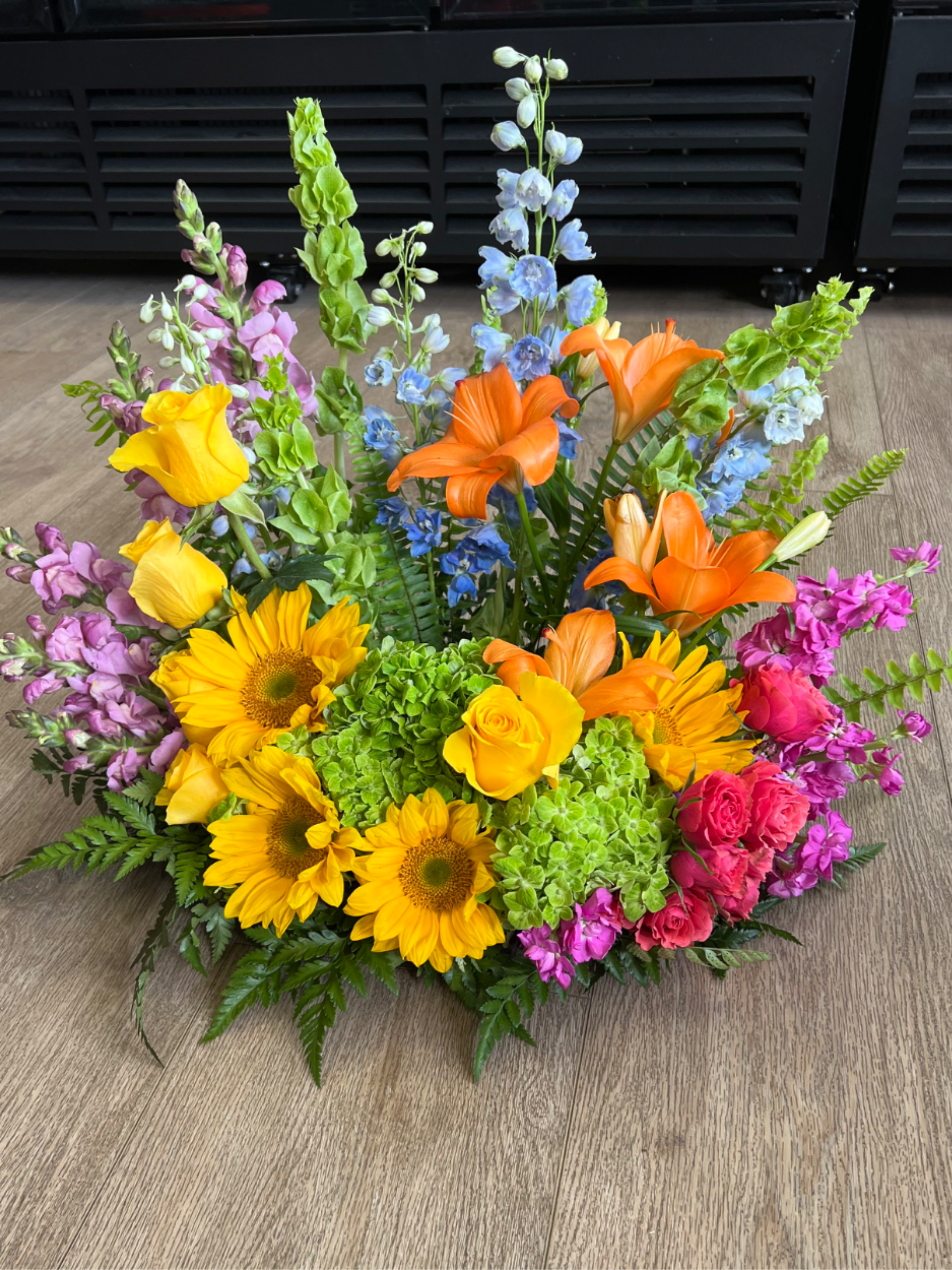 Low arrangement of yellow sunflowers, orange lilies, roses, and mixed bright blooms on a wood floor