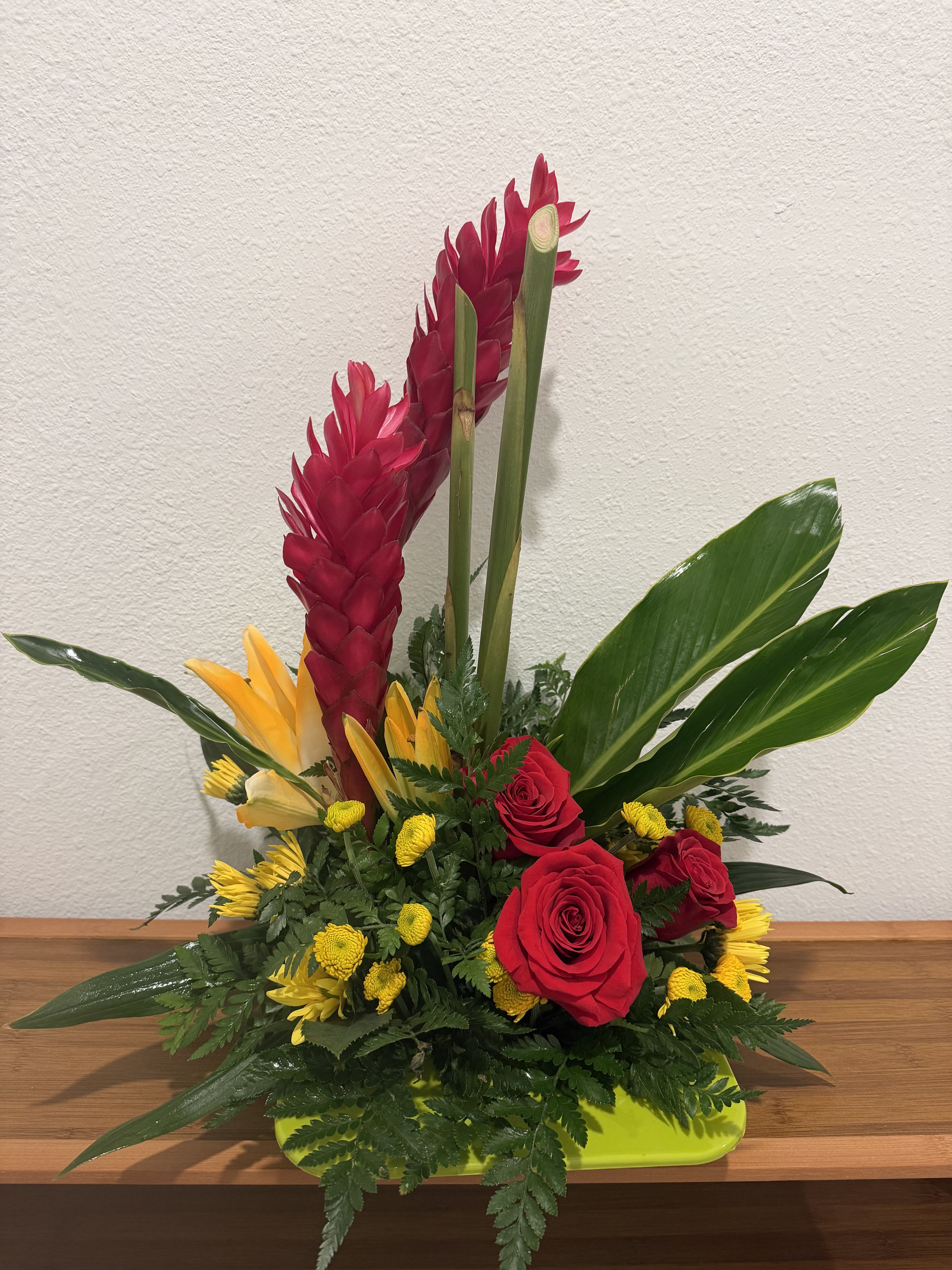 Tropical Passion Bloom - A vibrant tropical arrangement featuring bold red ginger, radiant red roses, yellow lilies, and cheerful yellow button mums accented with lush greenery. This eye-catching design brings warmth, energy, and exotic beauty to any occasion.