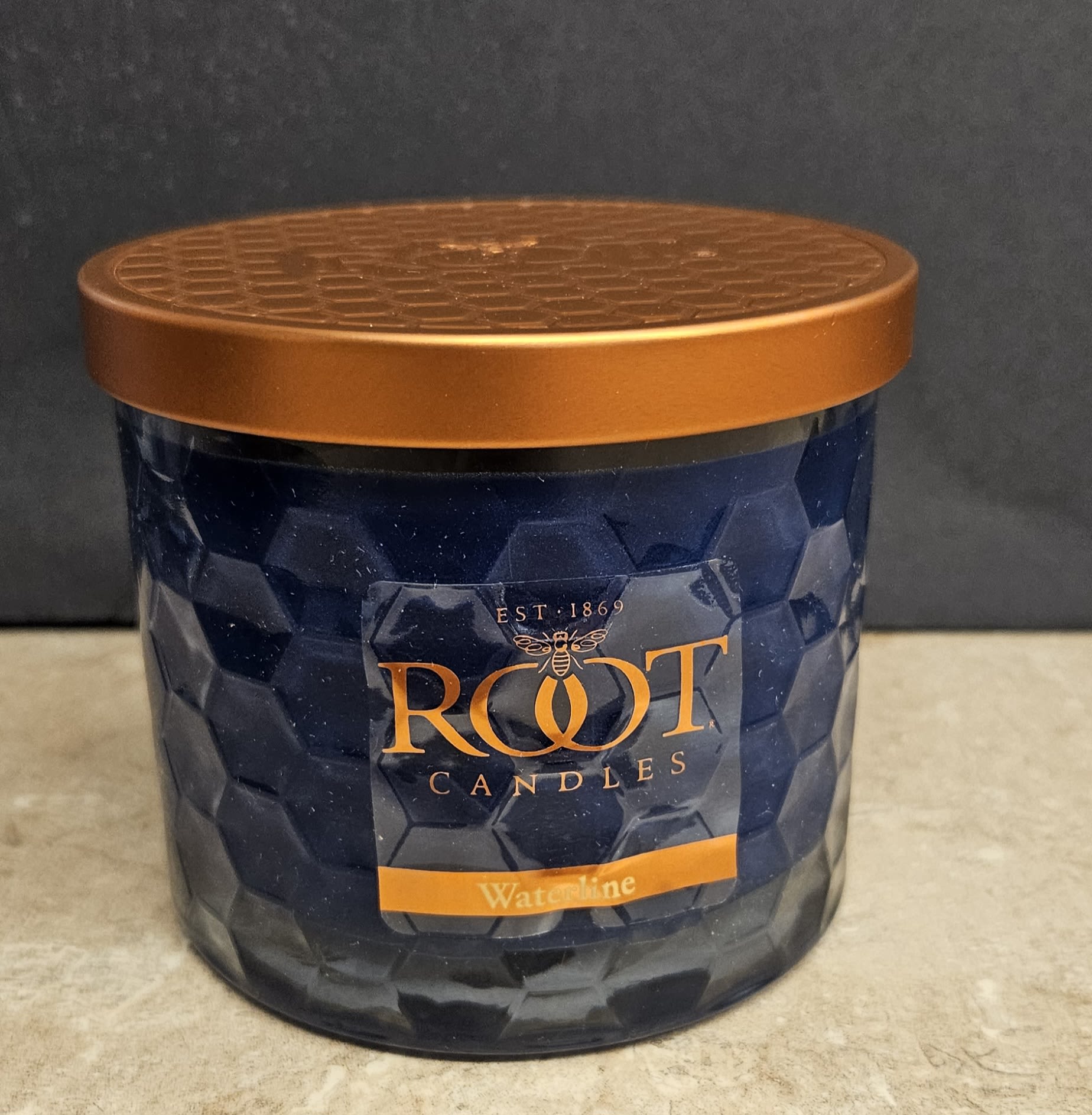Root Candle Waterline 3 Wick - Deep blue faceted glass meets warm copper in a moody, modern scented candle perfect for housewarming or everyday gifting. This Waterline candle reads like a thoughtful home accent-the dark blue container and metallic lid create a striking contrast that will catch the eye on a mantel, entry table, or bedside. Give the person you're thinking of a gift that elevates a room's atmosphere and signals care without being over the top.  The candle comes in a deep blue glass jar with a branded front label (Root Candles, Waterline) and a copper-colored metal lid, packaged as a ready-to-present home fragrance. Colors harmonize between blue, orange and black tones in the design, lending a contemporary, slightly moody look that suits modern interiors. Ideal for housewarming, thank-you, or "just because" moments-no fuss, just a polished scent and style that helps someone's space feel settled and seen.