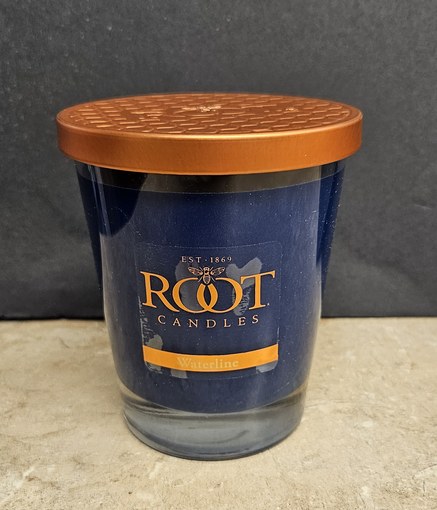 Root Candle Waterline L - Deep, calming navy wax topped with a copper-orange metal lid makes this single jar candle a striking gift for birthdays, housewarmings, or to say you're thinking of someone. As a home fragrance gift or a centerpiece for a cozy evening, this glass jar candle reads as both modern and substantial - the contrast of blue and warm orange conveys quiet warmth that will make the person you're sending it to feel comforted and well cared for.  The candle comes in a clear glass jar bearing the Root Candles and Waterline label for a retail-polished presentation, and is finished with a matching copper-orange metal lid. No floral elements are included; the simple, bold color pairing and branded look make it ideal for someone who prefers refined, minimal décor. Packaged as a single jar candle, it's ready to gift or display on a mantel, bedside table, or bathroom shelf.