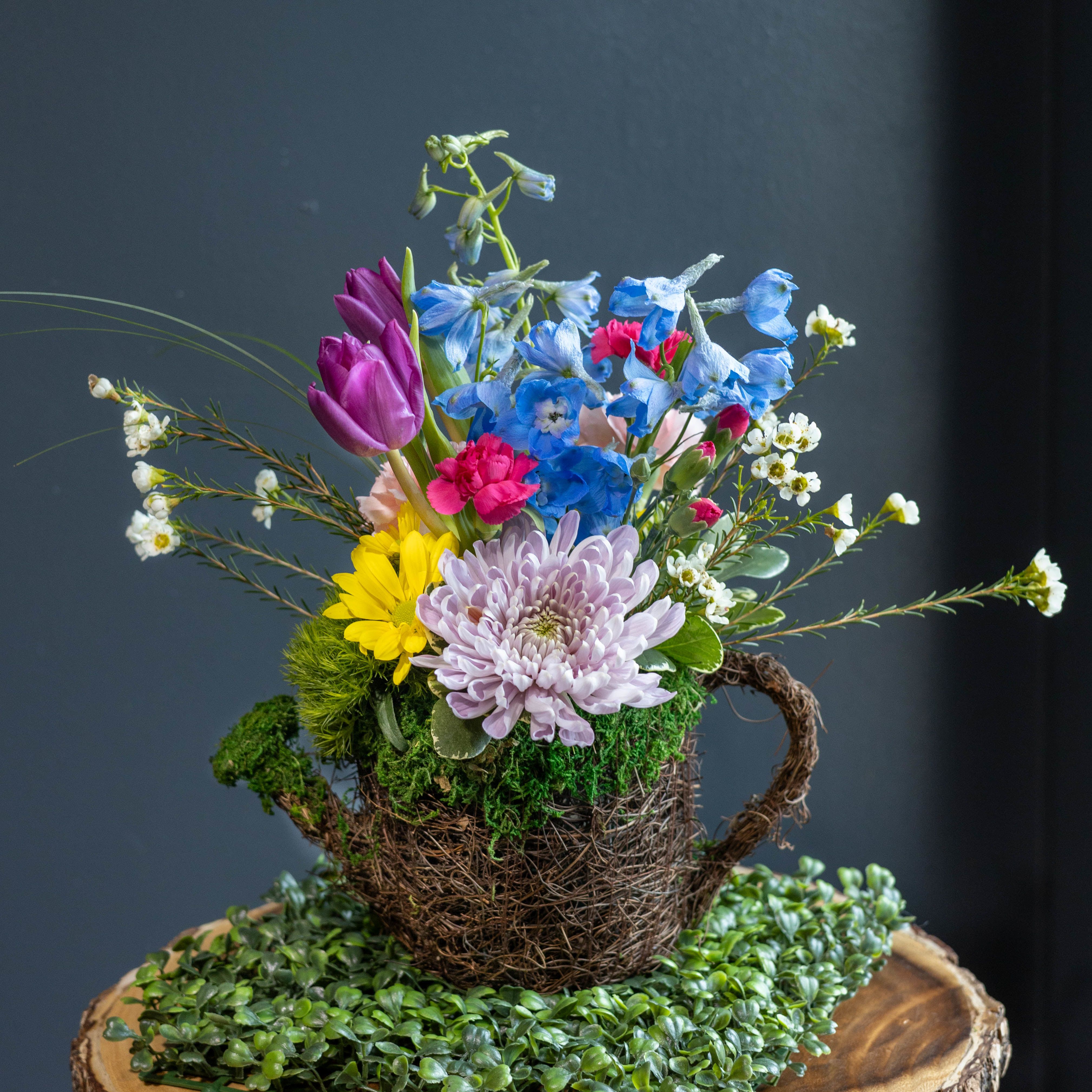Rainbow Garden - Celebrate the beauty of the season with Rainbow , a fresh and whimsical arrangement inspired by the gentle rains that bring spring gardens to life. Designed in a charming rustic watering can–style container wrapped in natural twig vine and soft moss, this arrangement captures the feeling of a blooming garden just after a spring rain.  A cheerful mix of seasonal flowers creates a vibrant palette of color and texture. Bright blue delphinium, soft lavender cushion mums, sunny yellow daisies, delicate pink carnations, and graceful purple tulips are artfully arranged with airy waxflower and fresh greenery. The playful design feels like a tiny garden in bloom, perfect for welcoming warmer days and brighter skies.  This delightful arrangement makes a wonderful gift for birthdays, spring celebrations, thank-you gestures, or simply to brighten someone’s day.