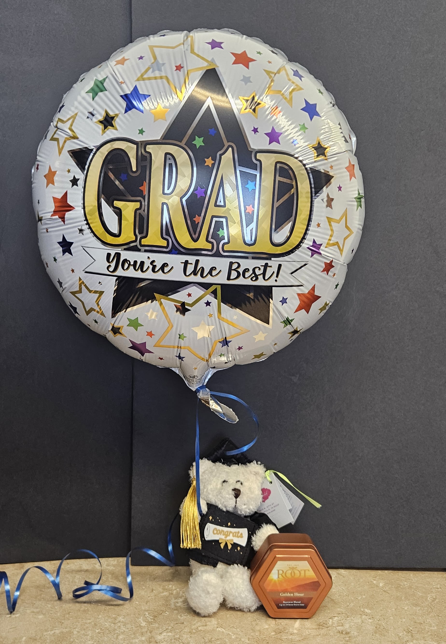 Graduation Bear with Balloon and Tin Candle - Celebrate their big achievement with a playful graduation gift set that puts the grad front and center. This flower arrangement-style presentation features a large round foil balloon printed with multicolored stars and bold "GRAD" and "You're the Best!" messaging, paired with a small teddy bear in a miniature graduation outfit complete with tassel and "congrats" label - a cheerful congratulations gift for graduation or a job milestone that will make the recipient feel recognized and proud.  Accenting the festive look are mixed-color accents with touches of white, black and orange in the styling, a hexagonal gift box for a small surprise, and ribbon and graduation-cap decoration to complete the theme. The included tin candle adds a cozy finishing touch, turning this into a ready-to-give bundle that looks lively on a dorm room dresser or office desk.  Perfect for the new grad, proud parent, or teacher you want to honor, this set is the kind of gift that makes someone feel truly celebrated and remembered. Easy to order and delivered assembled, it's a thoughtful, display-ready way to say congratulations-no extra wrapping required.