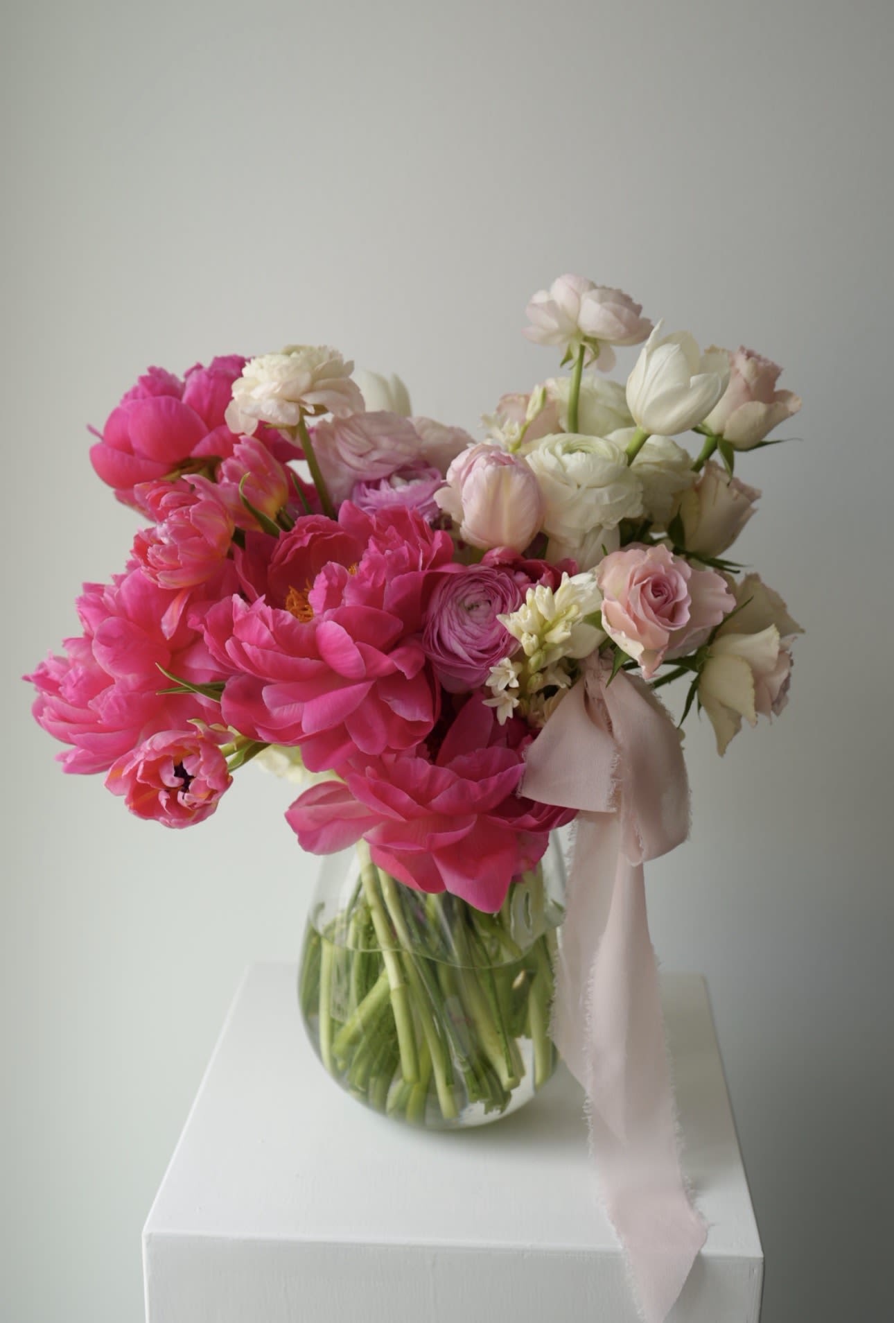 La Pivoine - A soft and romantic spring arrangement in layered shades of blush pink, ivory, and vibrant peony tones.  Designed with lush peonies, garden roses, ranunculus, tulips, and delicate seasonal textures in a clear glass vase finished with flowing silk ribbon.  Airy, elegant, and full of movement — like spring captured in bloom.