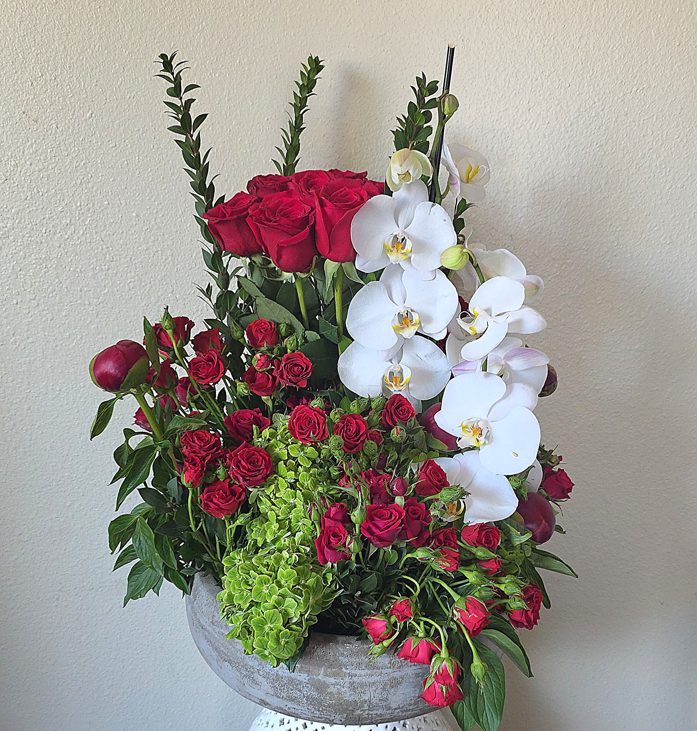 Exquisite Embrace - Deep clusters of deep red roses meet cascading white Phalaenopsis orchids in a dramatic anniversary bouquet built to make someone feel deeply cherished. This flower arrangement for anniversaries, romantic occasions, or refined everyday gifting uses strong color contrast-saturated red and crisp white-to create an immediate emotional impression: romantic, polished, and unmistakably special.  The design pairs lush red roses with bright green hydrangea-style blooms and upright greenery for texture and height, forming a layered, sculptural silhouette. White Phalaenopsis orchids trail gracefully for a soft, elegant contrast, and the whole composition is presented in a rounded gray ceramic bowl vase that grounds the arrangement with a modern, upscale feel. The asymmetrical shape and varied heights give it a statement-making presence ideal for a dining table, entryway, or bedside display.  Give this to the person you're thinking of when you want them to feel celebrated, adored, or seen-especially for anniversaries or moments of romance. The sturdy ceramic bowl keeps stems secure; to extend bloom life, refresh the water as needed and keep the arrangement out of direct heat and drafts.