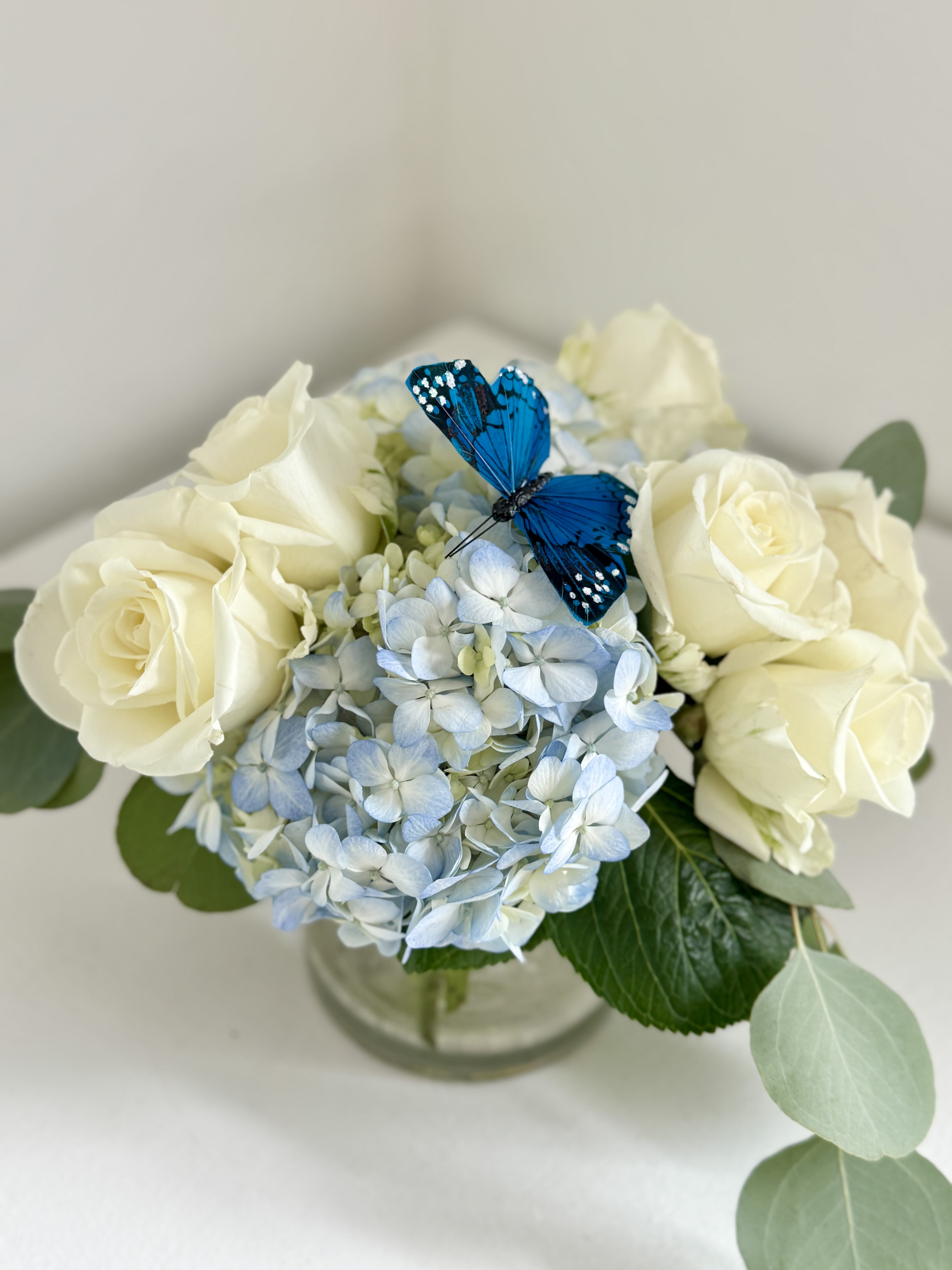 Cream roses and blue hydrangea in a glass vase with a blue butterfly accent