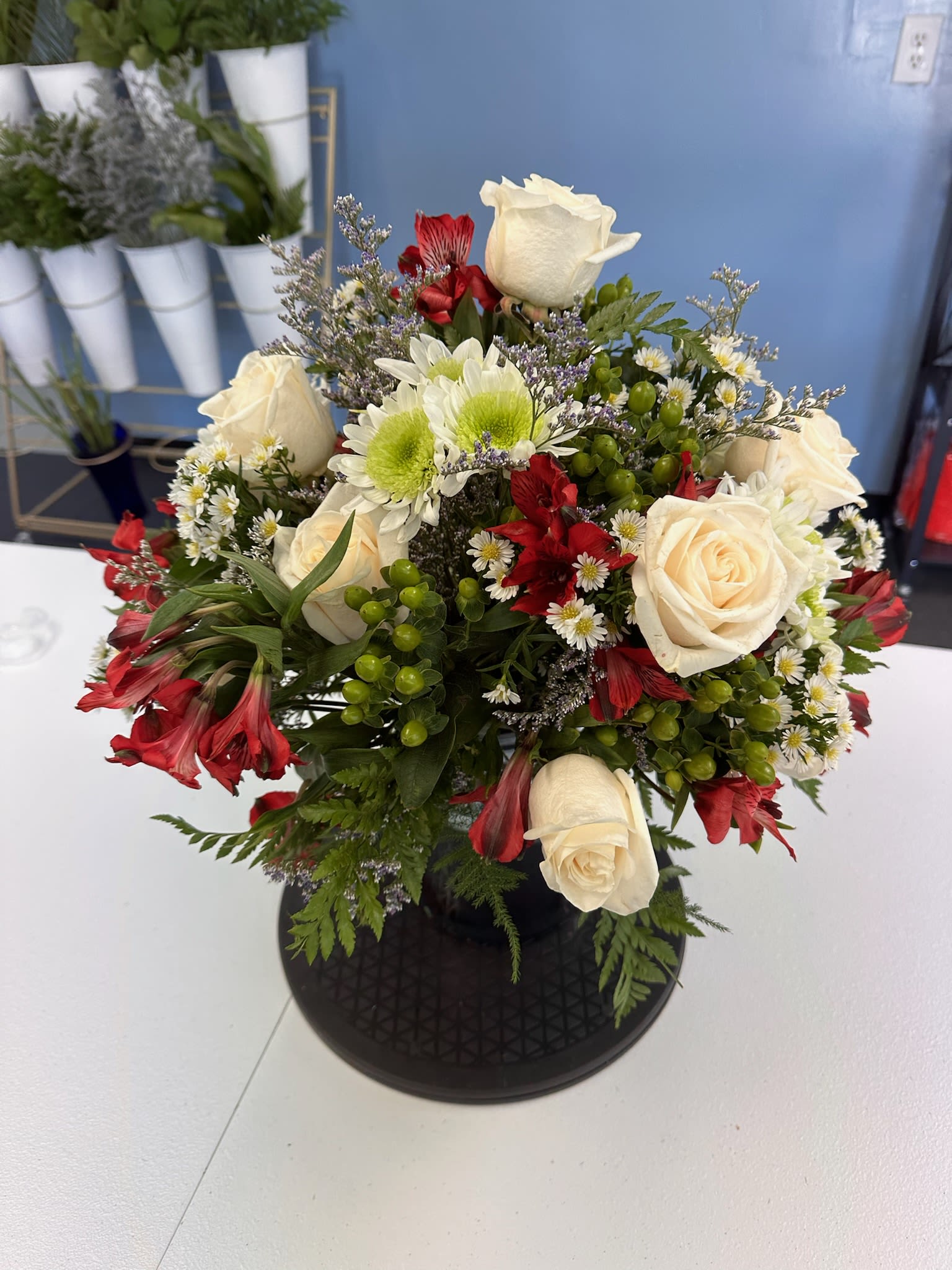 Celebration - Cream roses and vivid red alstroemeria take center stage in this flower arrangement for anniversaries, sympathy, or everyday gifting-an elegant celebration bouquet that reads classic and slightly formal. The mix of cream and red immediately signals warmth and affection, while the bright green berry clusters and airy purple accents give the person you're thinking of a lively, hopeful lift.  This rounded design pairs white daisy-like blooms and green button chrysanthemums with soft pink touches and small filler flowers for a textured, full look. Arranged in a polished black vase, the centerpiece feels substantial and modern; the contrasting colors-cream, red, white, green and hints of pink and purple-create visual movement and depth that will draw the eye from every angle.  Perfect for marking a meaningful moment or offering comfort, this bouquet makes someone feel seen and celebrated. The black vase provides a ready-to-display presentation; choose the size that fits the space and we'll deliver it arranged and water-ready so the recipient can enjoy the blooms right away.