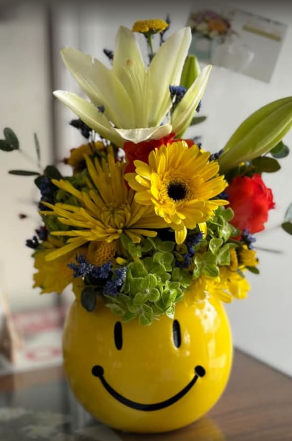 Happy Life - A glossy yellow ceramic smiley-face vase holds a cheerful mixed bouquet that instantly reads as a birthday bouquet or a just-because flower arrangement to lift someone's spirits. White lilies tower above sunny yellow gerbera daisies and warm chrysanthemums, while a single red accent bloom and sprays of small blue filler flowers add playful contrast and visual pop-perfect for someone who appreciates bold, happy color.  The composition feels upbeat and casual, designed to make the recipient feel seen and celebrated. Textures range from the lilies' smooth, fragrant petals to the daisy centers' sunny roundness and the chrysanthemums' full, cushiony heads. The arrangement arrives ready to display in the glossy ceramic vase; water and basic care-trimming stems and refreshing water every few days-will keep the flowers looking fresh through the week.
