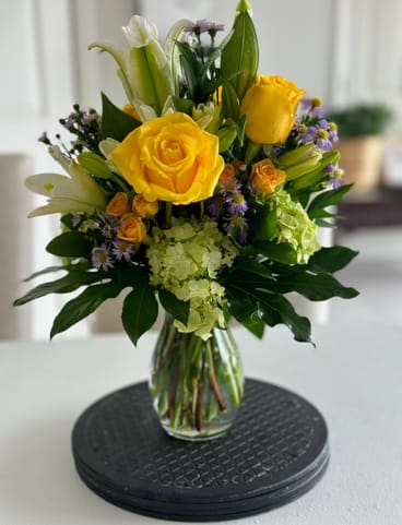 Josie Ann - Sunny yellow roses take center stage in this cheerful flower arrangement, making it an ideal birthday bouquet or congratulatory gift that will brighten someone's day. The mix of white lilies and pale green hydrangea clusters adds a soft, elegant contrast while small purple asters and spray roses create delicate, convivial accents-perfect for sending warm wishes or celebrating a special milestone.  The design is full and upright with a garden-style layering of rounded roses and hydrangea against the pointed shape of lily buds, giving the arrangement lively texture and movement. Arranged in a simple clear glass vase, the stems are visible for a fresh, natural look that suits both casual and more polished spaces. Colors include sunny yellow, crisp white, soft green, and touches of purple to keep the overall palette bright and balanced.  This bouquet is the kind of gift that makes the person you're thinking of feel noticed and celebrated-great for birthdays, get-well wishes, or "just because" surprises. The clear vase arrives ready to display; swap water and trim stems every few days to keep blooms looking their best.