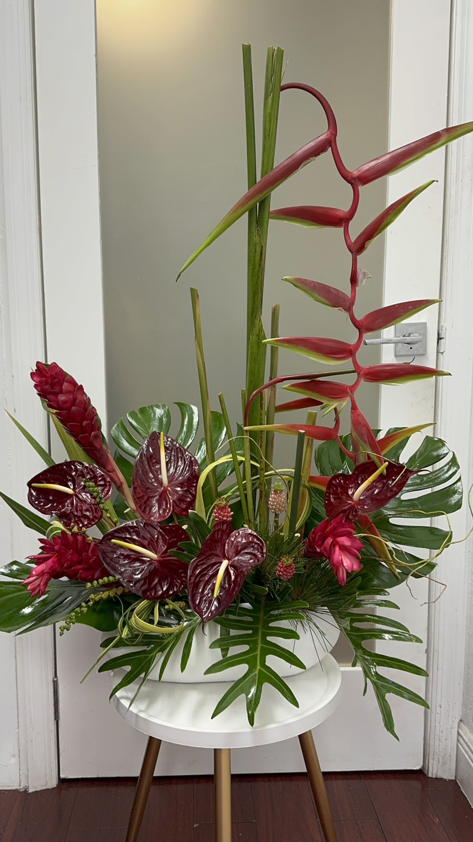 Tropical arrangement with red heliconia and burgundy anthuriums in a white container