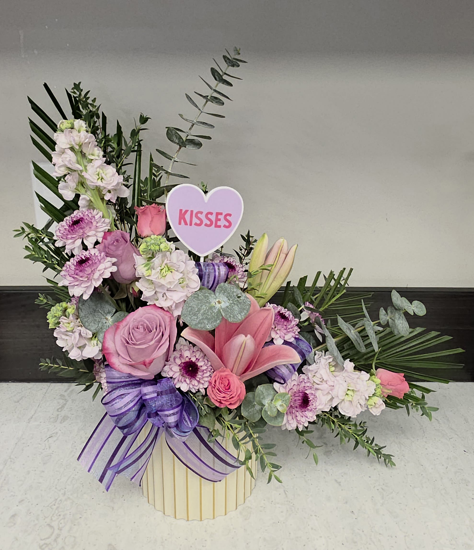 "L" for Love - A playful heart pick and lush pastel blooms make this "L" for Love bouquet a joyful choice for a romantic bouquet or a cheerful anniversary roses alternative. Bright pink and lavender roses mingle with pink lilies and pale stock to create a softly romantic first impression, while tropical leaves and silvery eucalyptus add texture and movement so the person you're thinking of feels truly celebrated the moment they see it.  Depending on the version you choose, the arrangement may arrive in a striped decorative box or a vase, finished with a satin or layered ribbon bow-options include purple ribbon with a "KISSES" heart pick for an affectionate touch, or layered blue-and-yellow bows for a fresh springtime look. Other variations feature pastel blue roses and tall white snapdragons for added height, or a dense, rounded cluster of hot-pink carnations and alstroemeria accented by bright green pom-poms for a bold, festive statement. Sizes are compact but full, designed to sit beautifully on a table or mantel and read as an immediate gift.  Perfect for anniversaries, birthdays, congratulations, or a bright just-because surprise, this arrangement sends a message of affection and celebration. Care is simple-keep blooms in fresh water, trim stems every few days, and place away from direct heat to prolong enjoyment; select your preferred container and ribbon style at checkout to customize the presentation.