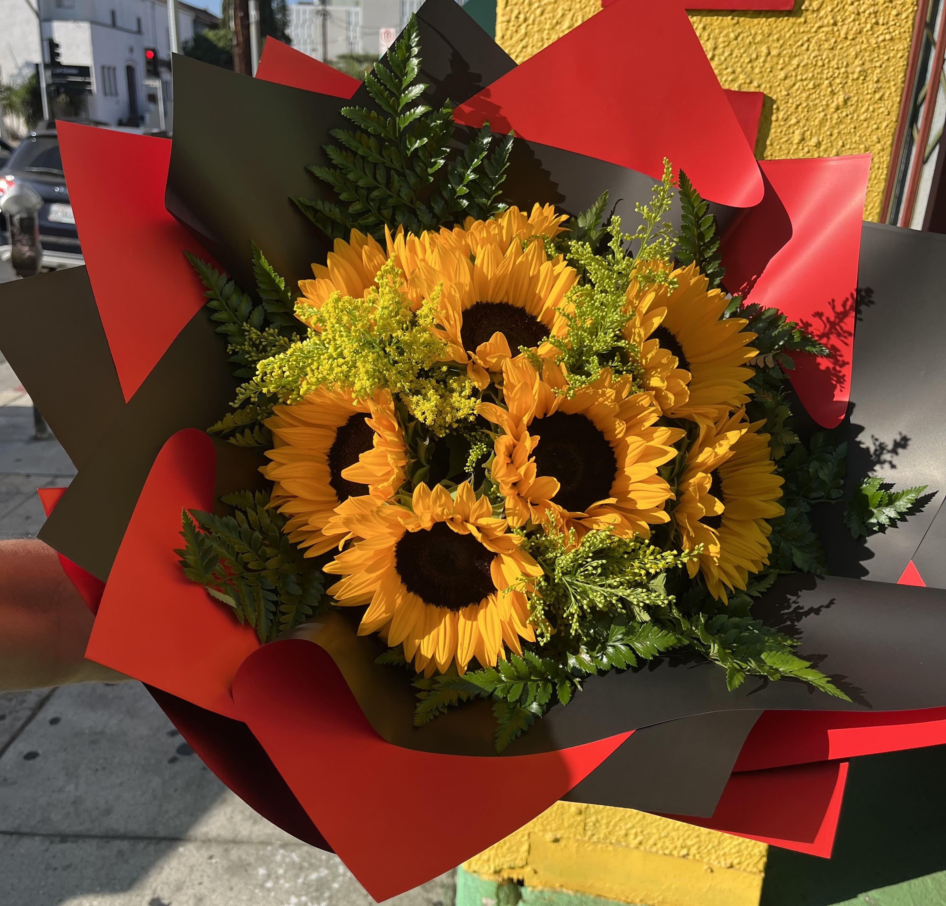 Hand-tied bouquet of yellow sunflowers with filler flowers wrapped in red and black paper.
