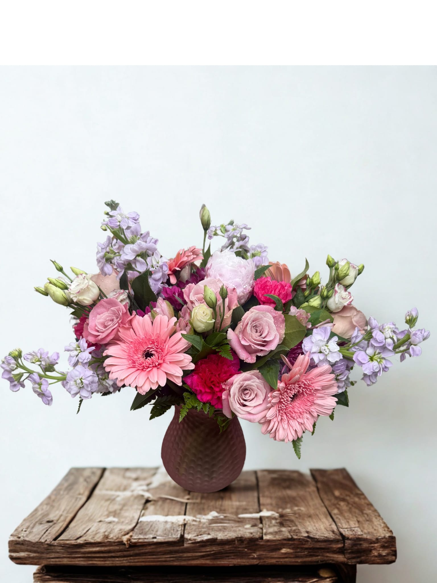 Blush Pink Boquet - A lush blush-pink bouquet that reads like a breath of spring-this flower arrangement is ideal as a birthday bouquet, anniversary gift, or a cheerful everyday surprise for the person you're thinking of. Pink roses and pink gerbera daisies give a romantic, uplifting focal point while pale lilac stock and soft lisianthus add delicate fragrance and layered texture; the mix of blush, magenta accents, white highlights, and fresh green foliage creates a joyful, celebratory feel.  Designed low and full, the arrangement is arranged in a textured dark ceramic vase that grounds the airy blooms with a modern, handcrafted presence-perfect for desks, dining tables, or bedside spaces. Carnations add volume and longevity while the stock brings subtle scent; the composition is balanced to read beautiful from every angle and to make someone feel truly seen on a special day or just because.