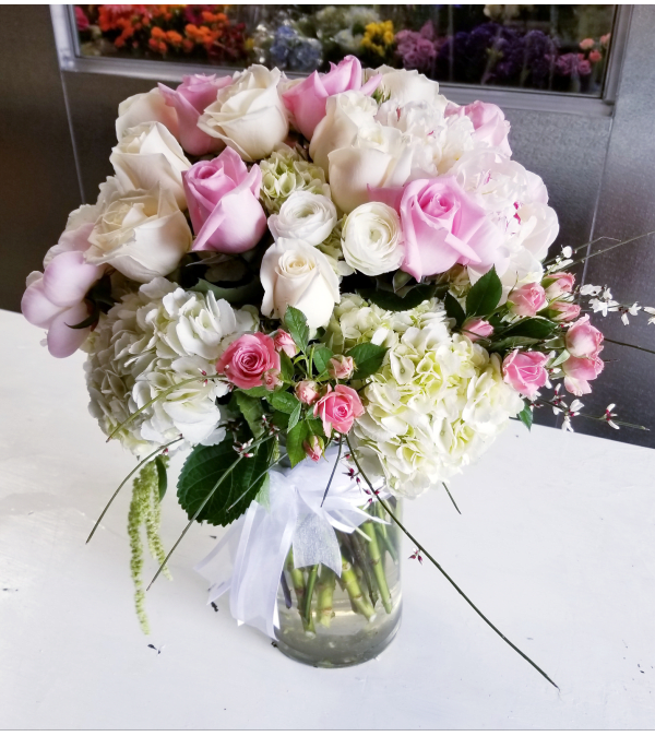 Round arrangement of white and blush pink roses with white hydrangeas in a clear glass vase with a white bow.
