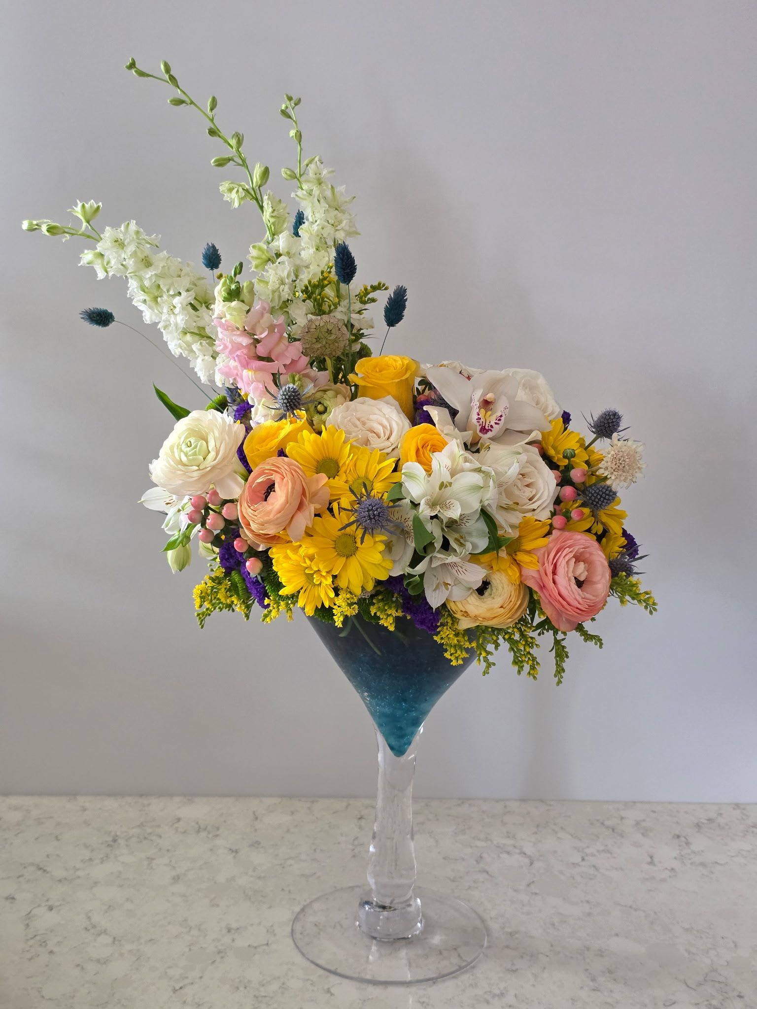 Golden Hour Martini Arrangement - Playful cocktail-glass arrangements that read like a tropical toast-perfect as a birthday bouquet or festive party centerpiece. These party-ready floral designs pair yellow roses and white daisies with green orchids and bright yellow accents in an oversized cocktail glass topped with a small blue paper umbrella, while the companion arrangement mixes cream roses, white daisies, blue blossoms and a fuzzy green pom-pom bloom in a purple-blue glass finished with yellow number toppers. The result is cheerful, whimsical, and unmistakably celebratory for birthdays or any festive gathering.  Each composition blends mixed roses, daisies, orchids, and complementary filler flowers in a low, wide cocktail-style vessel-one in clear glass with sunny yellow highlights and one in a purple-blue glass for contrast. Decorative hard goods-paper umbrellas, cocktail glasses, and number toppers-are included to amplify the party vibe. These compact arrangements are designed to sit on tables or be handed as a playful gift that makes the recipient feel singled out and celebrated; they arrive ready to display and enjoy with simple fresh-water care to keep blooms looking bright through the celebration.