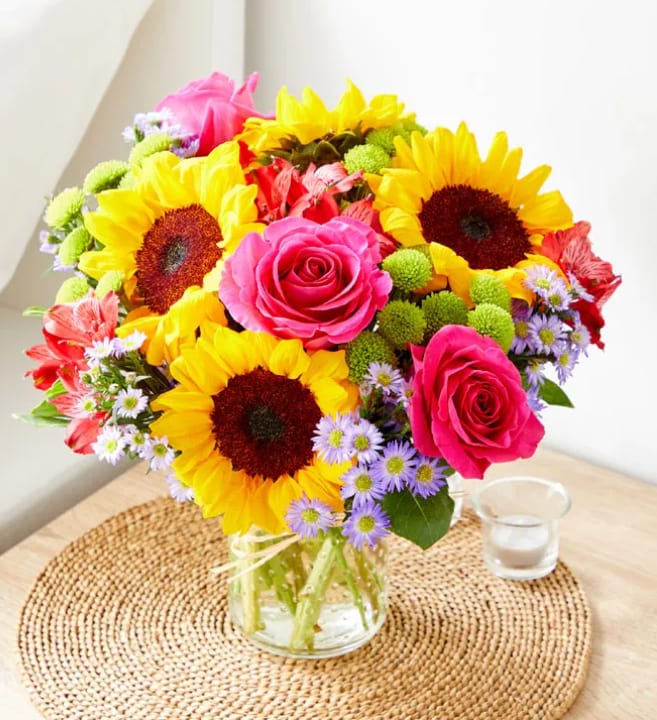 Fields of Gold Bouquet - Sunflowers take center stage in this Fields of Gold bouquet, a joyful flower arrangement perfect for birthdays, congratulations, or a bright "just because" surprise. The mix of sunflowers, vivid pink roses, and lavender daisy-like blooms creates a warm, celebratory mood-she'll feel noticed and cheered the moment the arrangement arrives.  Built as a full, rounded composition, the bouquet pairs warm yellows and pinks with cooler purple accents for lively contrast. Alstroemeria adds delicate texture alongside the daisies and roses, and the stems are presented in a clear glass vase tied with a light ribbon for a fresh, casual tabletop look. Comes styled on a woven placemat with a coordinating glass candle holder in the image-an easy centerpiece that makes any room feel sunnier and more inviting.