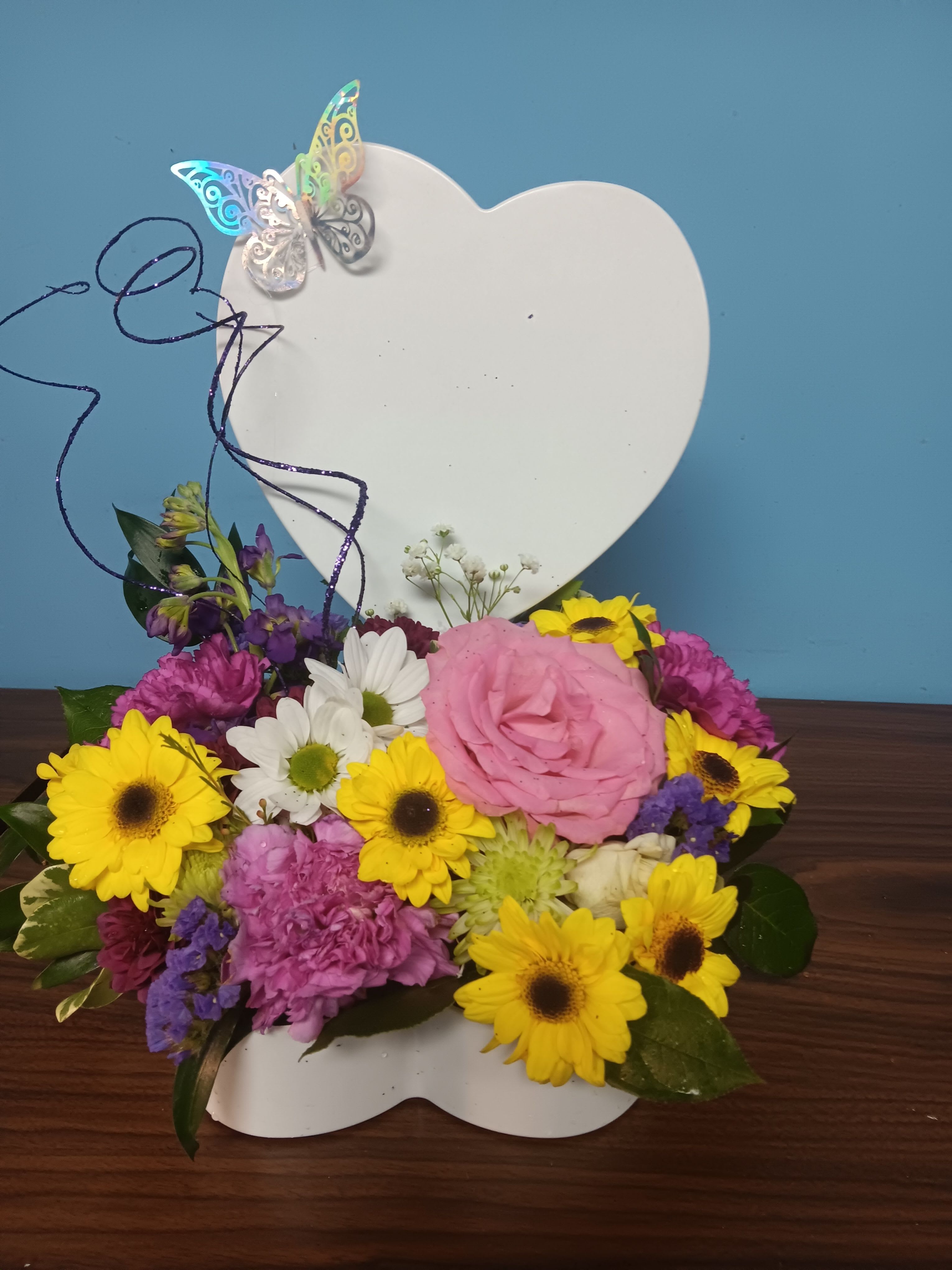 Sweetheart - Creamy roses and soft lavender blooms nestle against a white heart-shaped plaque in this compact Mother's Day flower arrangement, an ideal gift to make the person you're thinking of feel cherished and seen. The bouquet reads like a warm embrace - peach and white petals soften the palette while bright yellow accents lift the mood, so it works just as beautifully for an anniversary, a thank-you, or any sentimental moment.  This low, wide design layers roses, carnations, and daisy-like flowers to create a textured, romantic look with playful pops of color. A delicate decorative butterfly pick adds a whimsical touch to the heart backdrop, making the arrangement feel personal and keepsake-worthy. Perfect for tabletops or a bedside table, it arrives ready to display and brings a fresh, fragrant presence to the room.