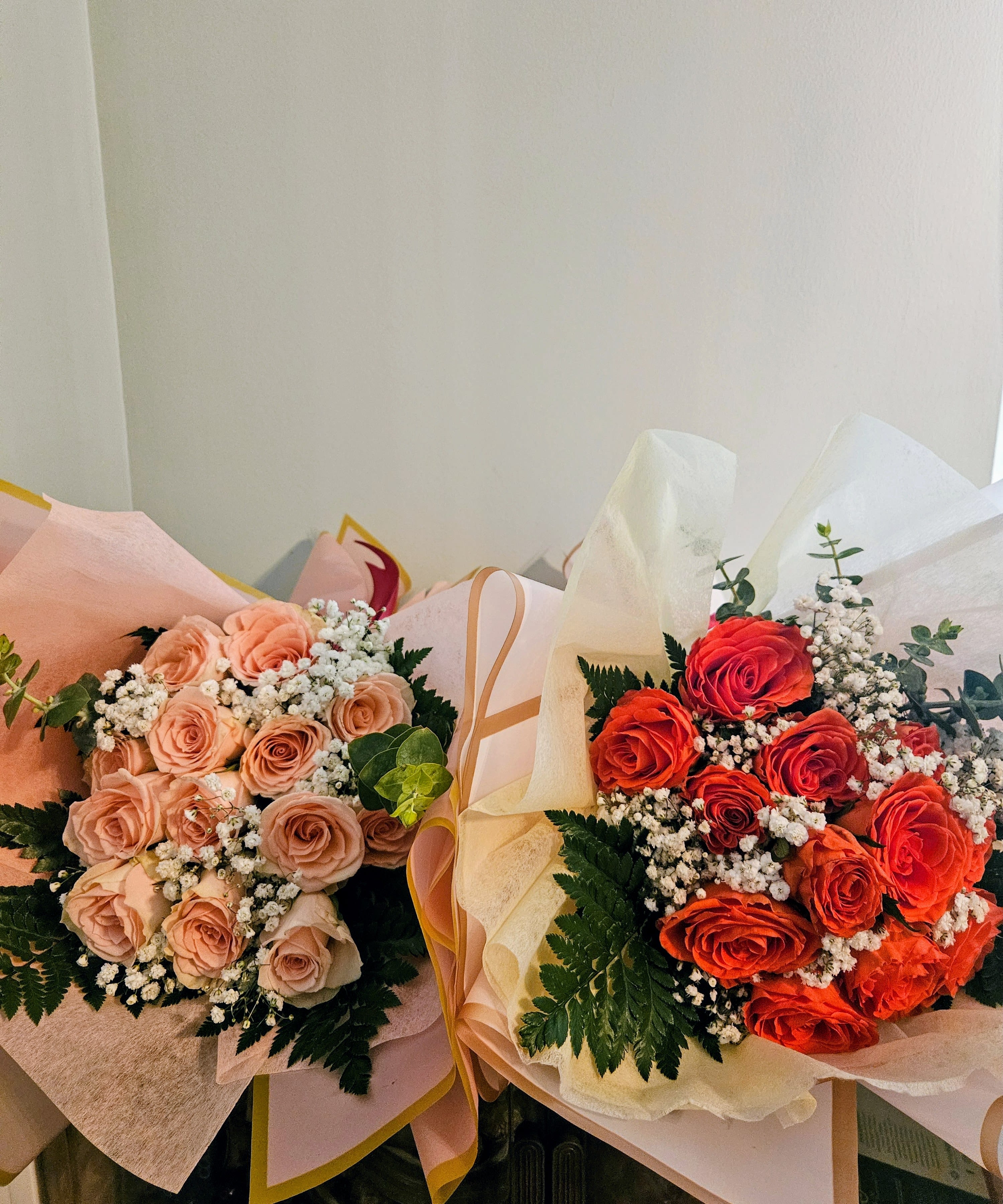 "Rose Bouquet" - Two hand-tied rose bouquets that read like a celebration: one in soft blush, peach, and white for a tender birthday or anniversary sentiment, and one in vivid red-orange and white for a bold romantic gesture. These rose bouquets are designed to make the person you're thinking of feel seen-whether you want them to smile with quiet warmth or flush with excitement. Perfect for birthdays, anniversaries, or just-because moments, each style sends a clear message of affection.  Each bouquet is built around clustered roses accented with small white filler blooms and rich dark green foliage for a full, rounded silhouette. The pastel-wrapped bouquet uses soft tissue paper and delicate wrap for an elegant, gift-ready presentation; the brighter bouquet is wrapped similarly but reads more energetic and passionate. Both are hand-tied for a natural, comfortable hold and arrive ready to display or hand to the recipient.  Choose the softer palette to convey gentle appreciation or the vivid red-orange arrangement to express romantic fervor; either option creates that lift of surprise and warmth when presented. Bouquets come wrapped in coordinating paper with tissue for protection-simple fresh-care tips included at delivery to help the roses last longer so your gift keeps giving.