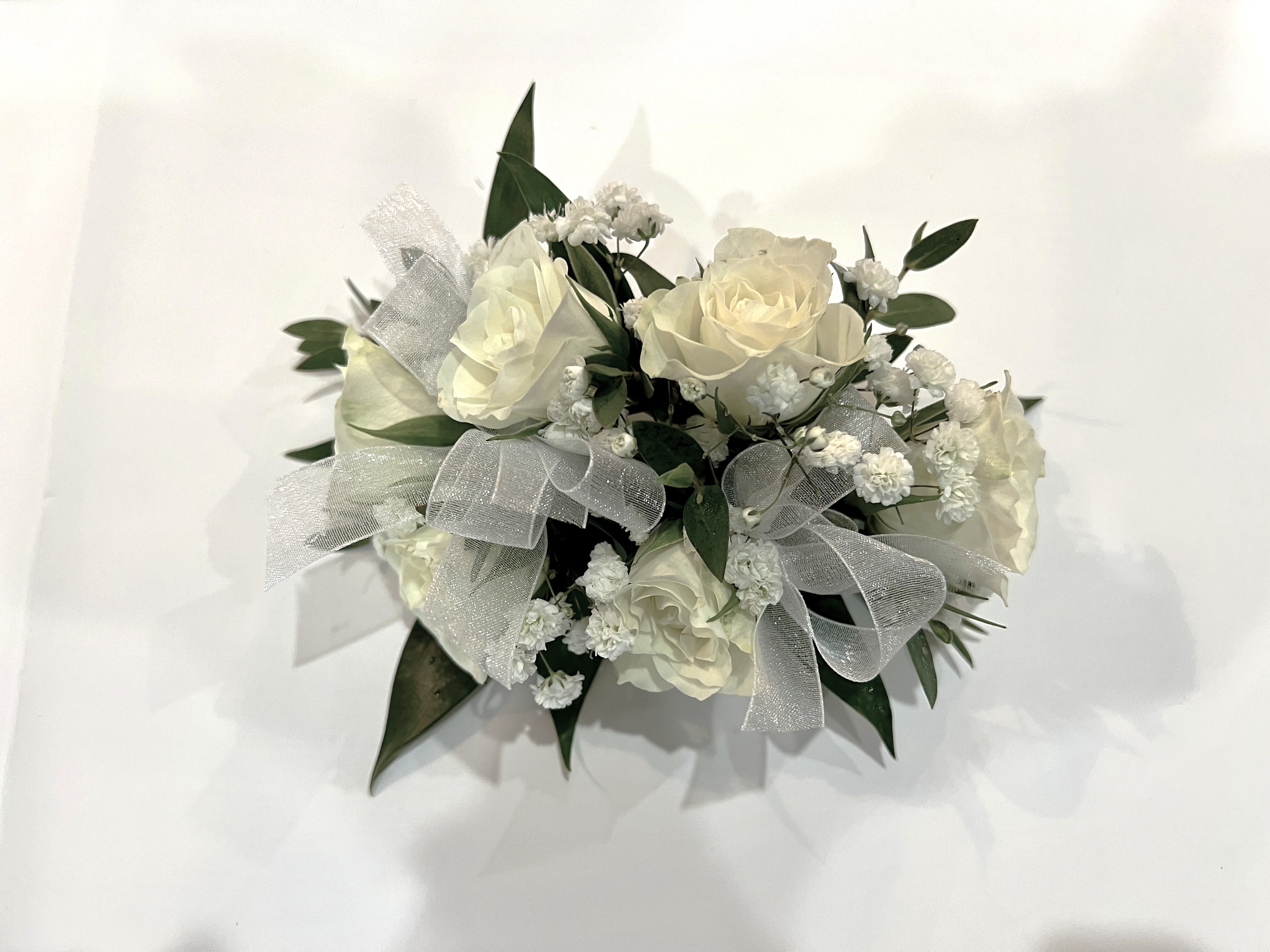 White Rose Corsage  - Creamy white roses gathered into a compact corsage create an elegant, romantic accent for weddings, sympathy, or a thoughtful everyday gift. The white rose corsage presents a restrained palette of white and deep green that feels fresh and formal-perfect for the person you're honoring on their wedding day, marking a solemn moment, or simply letting someone know you're thinking of them.  This low, rounded arrangement pairs clustered white roses with small white filler blooms and dark green foliage for a lush, tactile look. Wide white mesh ribbon is woven through the design, adding a soft metallic sheen and refined texture that complements formal attire. The corsage is compact enough to wear comfortably while still making a graceful statement.  Choose this corsage when you want the recipient to feel quietly celebrated-won't overwhelm an ensemble yet reads as thoughtfully chosen. Because the composition is compact and rounded, it sits neatly on a wrist or pinned to clothing; handle gently and keep cool to prolong freshness.