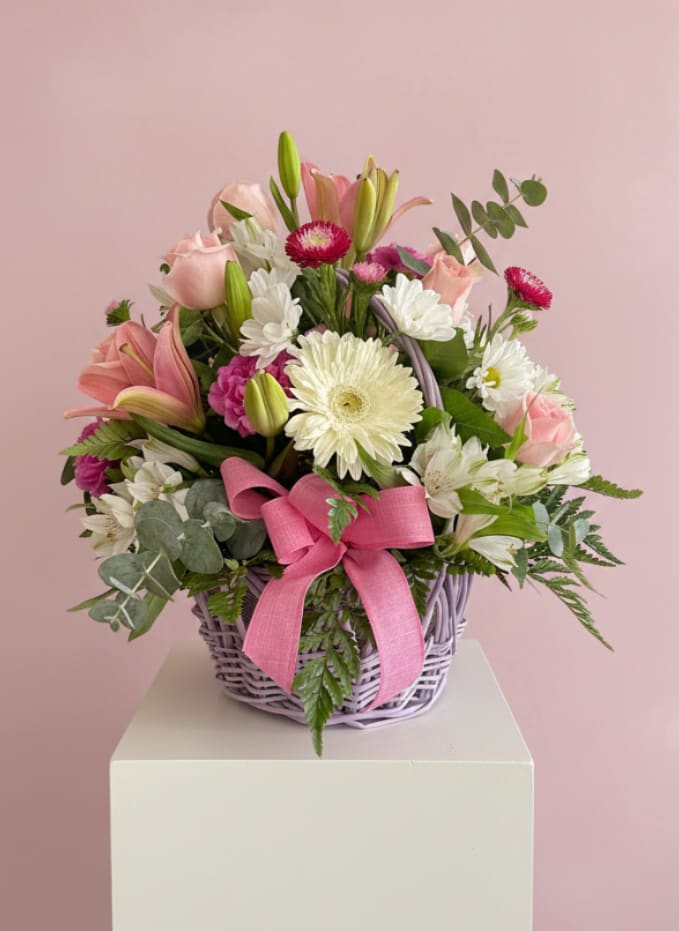 Gift to Exist - Pale pink roses and lilies mingle with creamy white gerbera daisies, chrysanthemums, carnations and alstroemeria in a rounded lavender wicker basket-a soft, romantic flower arrangement for a birthday, a "just because" surprise, or a spring thank-you. The few upright lily buds add graceful height while eucalyptus and fresh greenery soften the edges, creating a gift-ready presentation that feels like a gentle, celebratory embrace for the person you're thinking of.  Hand-layered for a lush, full look, the design is tied with a wide pink ribbon and arranged so each bloom shows its best side-romantic pinks, crisp whites, and touches of green and purple work together to make her feel seen and cherished. Because it's a gift to exist and a wonder to taste, this arrangement brings warmth and delight to the moment. For best enjoyment, refresh the water daily and, if you move the flowers to a vase, trim the stems to extend their life.