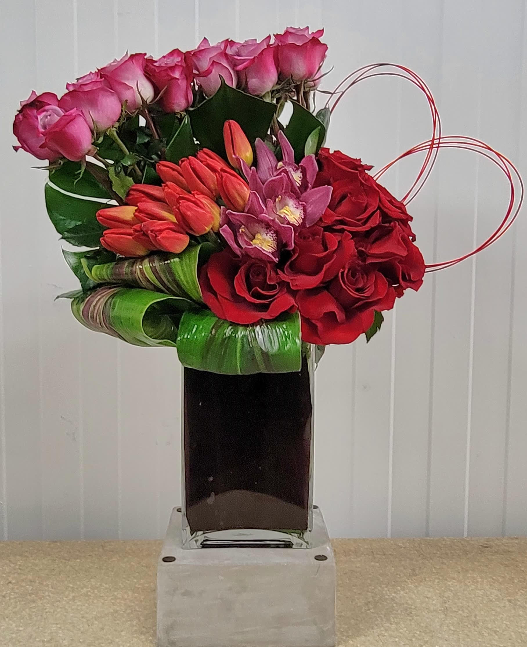Pink and red floral arrangement in a tall glass vase with heart-shaped wire accents
