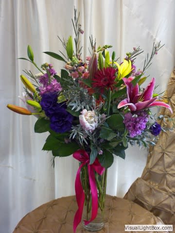 Mixed bouquet with lilies, purple blooms, and a pink ribbon in a glass vase
