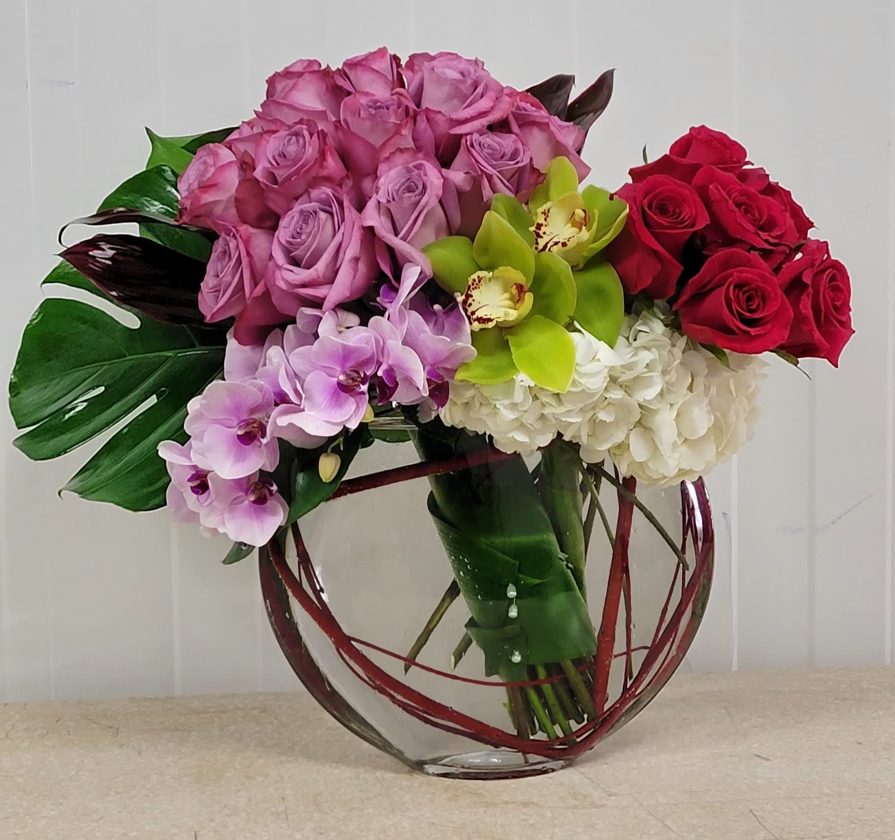 Low glass vase arrangement with lavender roses, red roses, green orchids, white hydrangeas, and purple orchids.