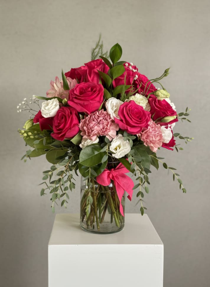 Spicy Mama - Lush hot pink roses take center stage in this Spicy Mama flower arrangement, a vibrant birthday bouquet or Mother's Day gift designed to make the person you're celebrating feel adored and seen. The vivid pinks paired with soft whites read as playful and heartfelt-perfect for anniversaries, Valentine's Day, or any moment you want to say "you brighten my life" with confident color.  Soft pink carnations, white lisianthus and alstroemeria, plus delicate filler blooms and airy greenery form a rounded, garden-style shape with gentle movement at the edges. Trailing foliage softens the silhouette and adds texture, while the clear glass vase and bright pink ribbon give the arrangement a polished, gift-ready finish that looks beautiful on a dining table, bedside, or entry console.  Made for the partner, mother, or close friend you want to celebrate, Spicy Mama feels both affectionate and festive-she'll feel genuinely seen when this arrives. Vase included; to keep blooms fresh, refresh the water daily, recut stems as needed, and display away from direct heat or drafts.