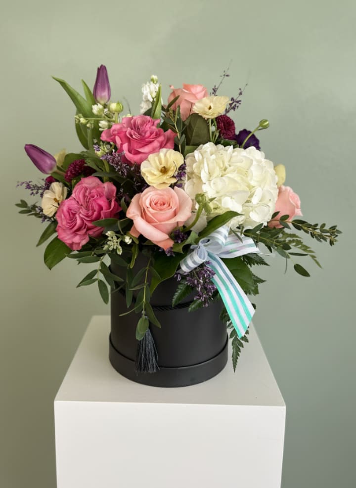 Feeling Beautified - A lush white hydrangea crowns a compact black hatbox surrounded by blush roses, deep plum tulips, and delicate dianthus. Feeling Beautified is a romantic flower arrangement that reads both soft and dramatic. As an anniversary bouquet or birthday gift, this layered palette of pink, purple, white, and fresh green foliage creates an immediate sense of warmth and thoughtful luxury for the person you're celebrating.  Arranged tightly in a chic black hatbox with a striped decorative ribbon, the design presents beautifully as a ready-to-give keepsake centerpiece. Flowers include hydrangeas, roses, tulips, dianthus, and small accent blooms; the mix of cream, blush, and rich plum tones ensures visual depth and texture from every angle. Sized as a compact hatbox arrangement, it arrives polished and gift-ready-perfect for saying "I love you," "happy birthday," or "you're on my mind" with an elegant, unforgettable flourish.