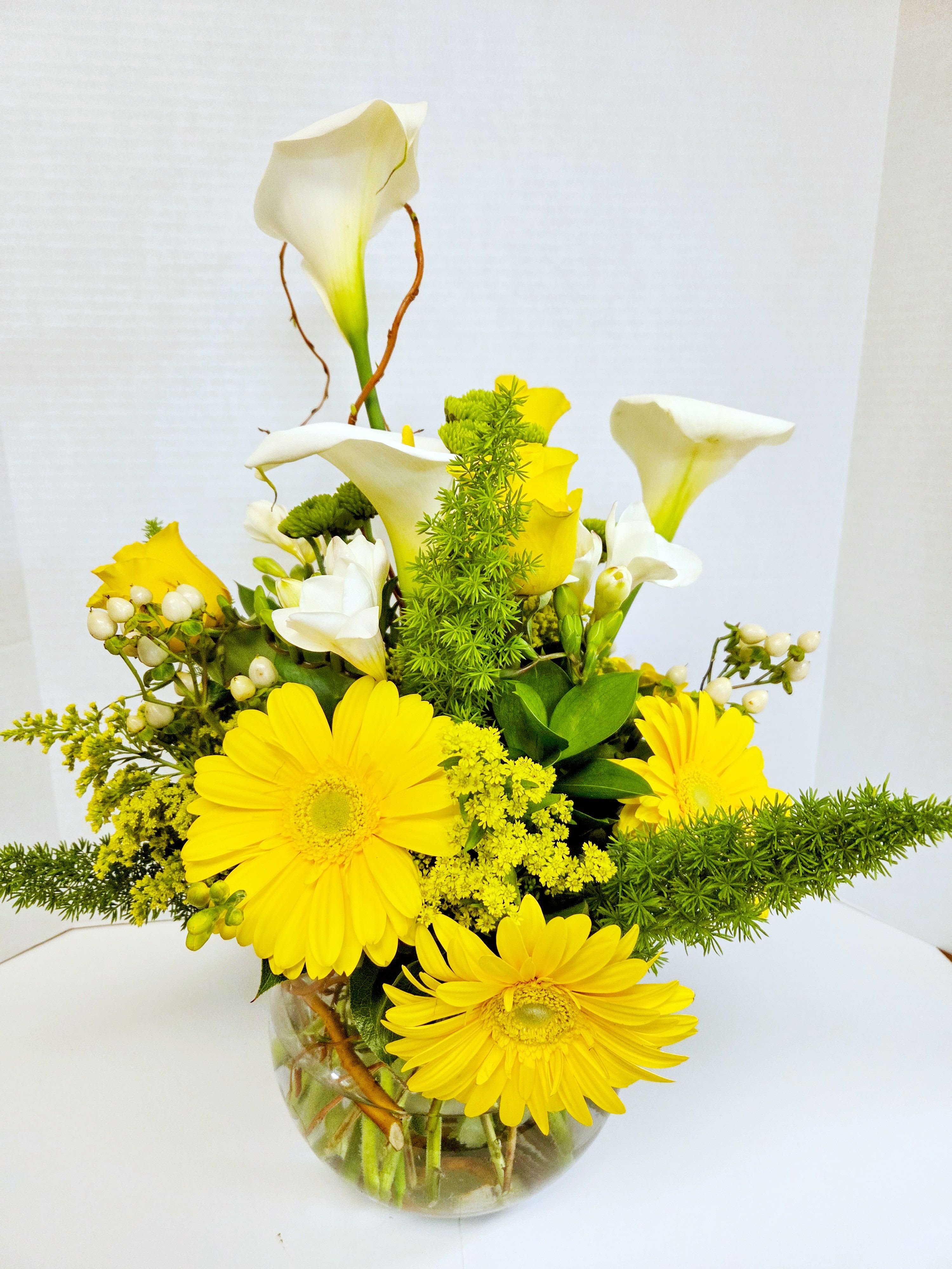 Compact arrangement of yellow gerbera daisies and white calla lilies in a clear round vase