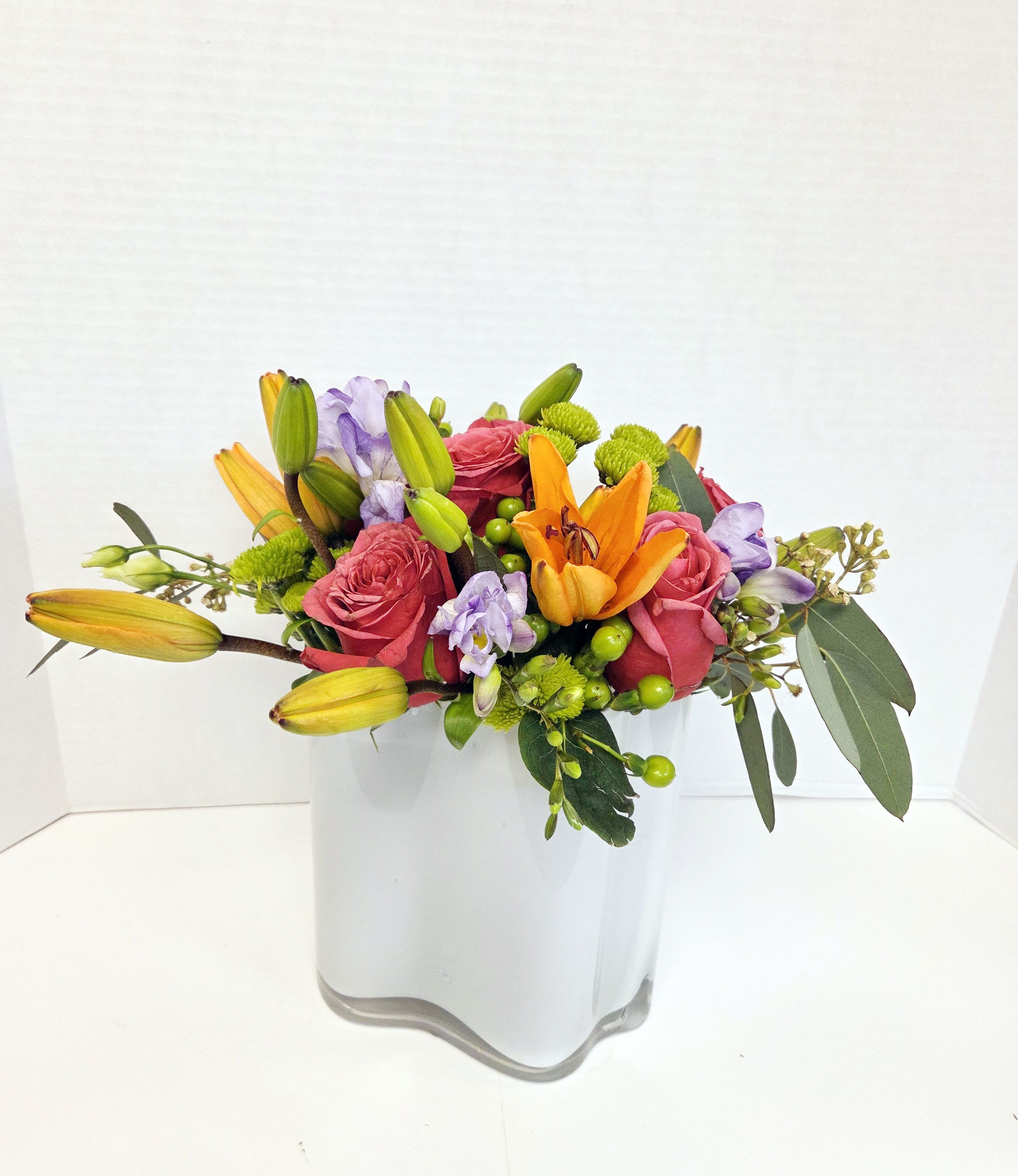Mixed bouquet of pink roses, orange lilies, and purple flowers in a white vase
