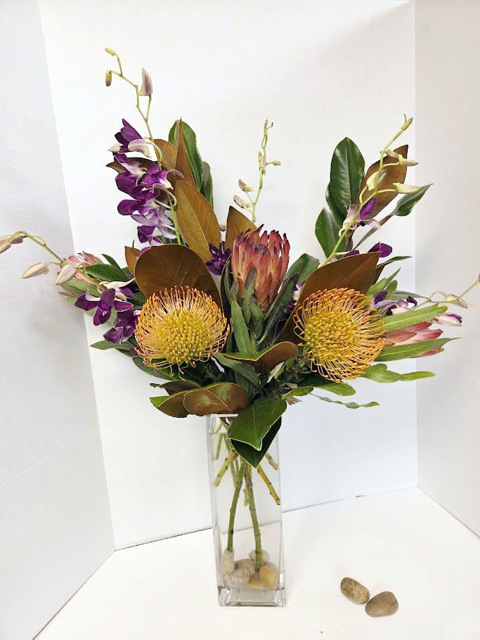 Tropical flower arrangement in a tall clear vase with purple orchids and yellow pincushion blooms