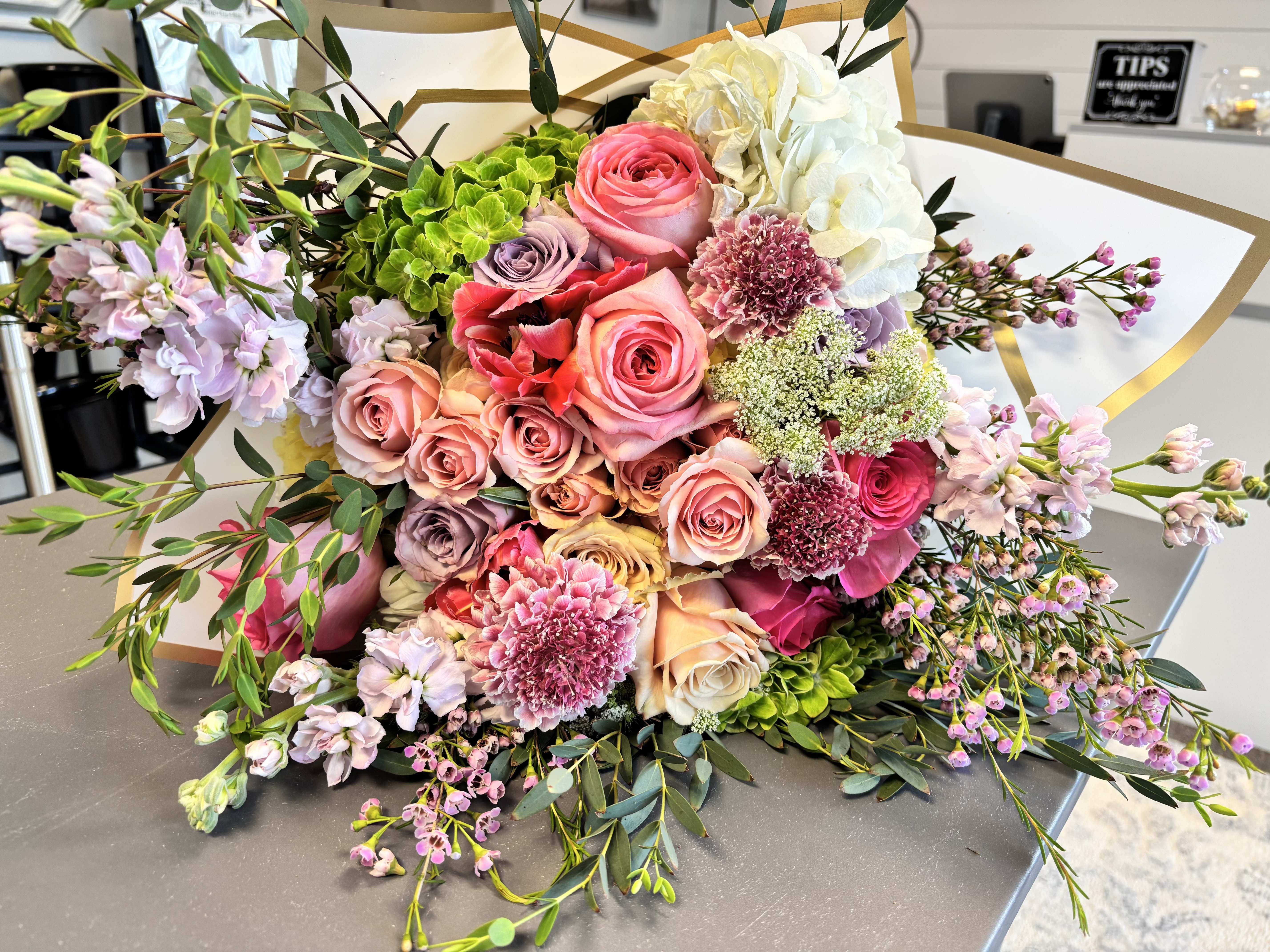 Sunshine Into the Next Chapter (Wrapped Bouquet) - A lush, garden-style bouquet overflowing with soft pastels-Wrapped in Sunshine is a large mixed bouquet perfect for an anniversary bouquet, birthday gift, wedding decor, or a heartfelt thank-you. Clustered roses in pink, blush, peach, cream, and lavender sit at the center, creating an immediate sense of romance and celebration that will make the person you're thinking of feel truly seen.  White and bright green hydrangea add volume and cloudlike texture at the top, while dianthus-like blooms, small pink sprays, and airy greenery spill outward for a generously hand-tied, slightly cascading silhouette. The arrangement is finished in decorative wrapping paper and tied with a gold ribbon for an elegant, gift-ready presentation-soft, feminine colors (pink, white, purple, green) combine for a joyful, sophisticated look.  Ideal for marking special moments or showing appreciation, this bouquet reads as both festive and intimate-the kind of gift that brightens a table and lingers in memory. Available in a large size as shown; to keep blooms fresh, trim stems and change vase water every 2-3 days and avoid direct heat or drafts.  A Ray of Sunshine Floral proudly offers graduation flower delivery in Hilliard, Dublin, Upper Arlington, Grandview Heights, and surrounding Columbus communities.