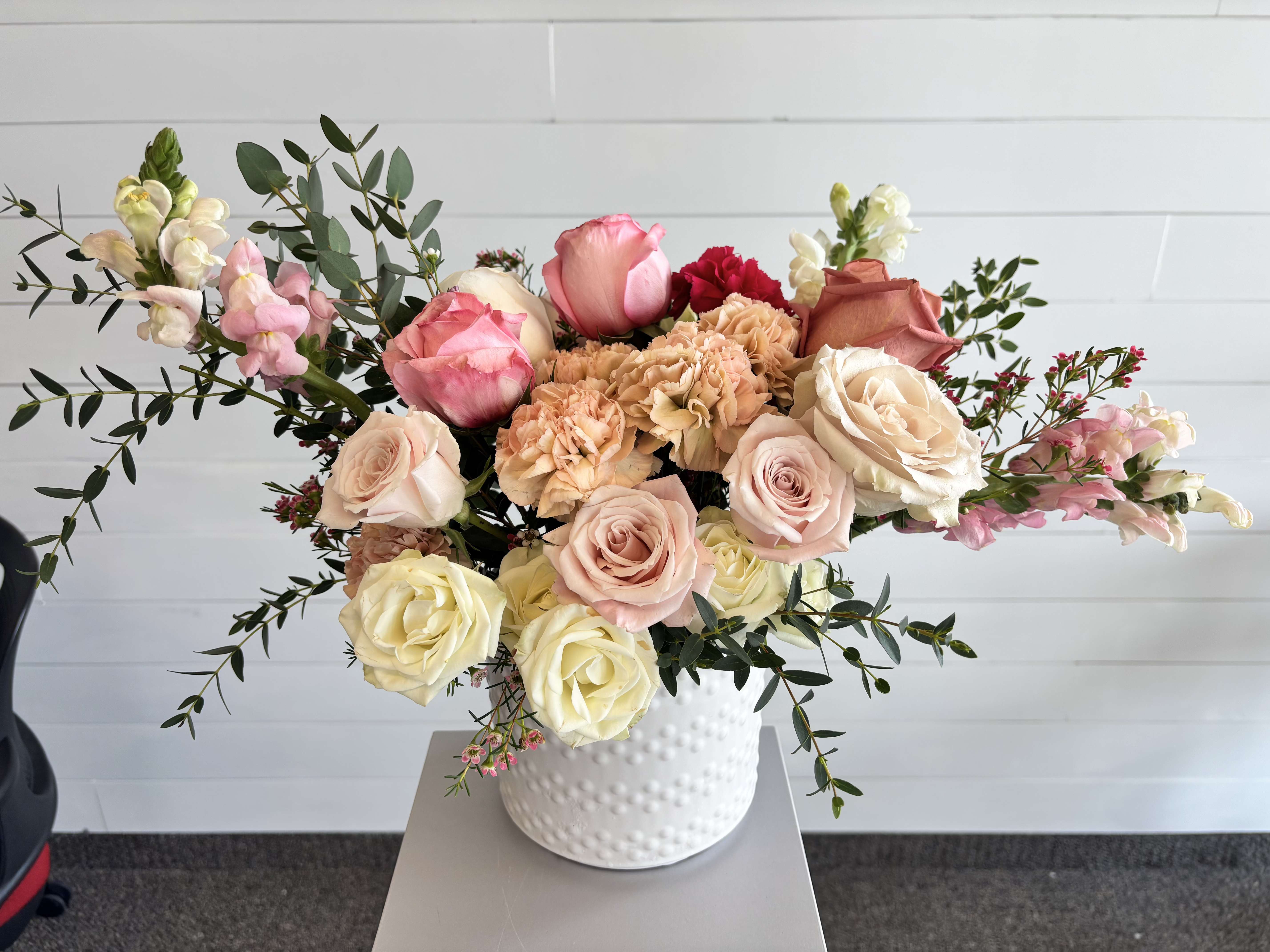 The Vicki (Vase Arrangement) - A soft, elegant garden-style arrangement designed with a refined, airy feel. Featuring a harmonious blend of blush, peach, and creamy white blooms with delicate accents and natural greenery, arranged in a textured white ceramic vase for a timeless, elevated look.  Includes:  Roses in soft blush, peach, and ivory tones Carnations and complementary seasonal blooms Accent florals for texture and movement Fresh greenery White ceramic vase  Each arrangement is hand-designed, with slight variations based on the freshest flowers available.