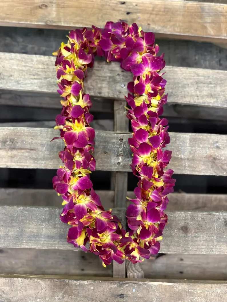 Double Leis - Sunset - A lush pair of double leis crafted from densely strung purple orchids with bright yellow centers creates an immediate island-ready impression-perfect as a graduation lei or a cheerful everyday gift. This flower arrangement for celebrations and casual gifting reads vibrant and festive; the magenta-purple petals against warm yellow accents make the recipient feel honored and joyfully seen.  Each lei is a thick U-shaped garland of orchids designed to drape comfortably around the shoulders or be displayed on a mantel. Shown against a weathered wooden bench for a rustic, tropical presentation, the arrangement highlights the orchids' velvety texture and vivid color contrast. Ideal for graduations, welcome-home moments, or any occasion that calls for a celebratory, sun-soaked touch-order ready-to-wear leis for easy gifting and lasting visual impact.