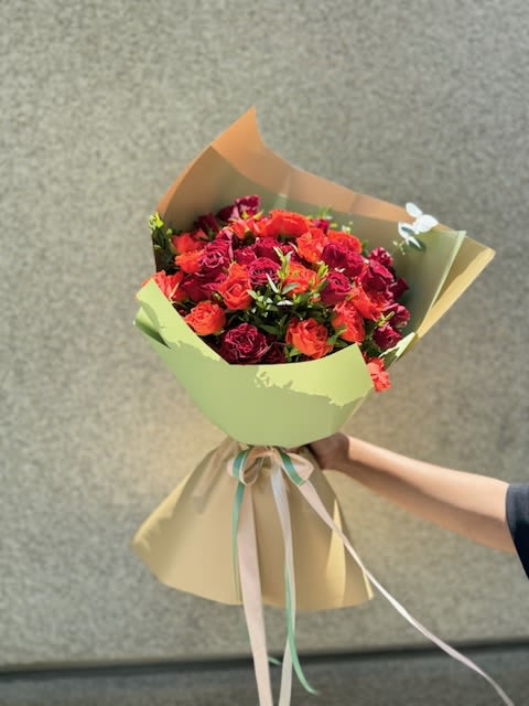 Spray Roses bouquet  - A lush cluster of spray roses in saturated reds, coral and deep pinks creates an immediate warm, romantic impact-perfect as a birthday bouquet, anniversary surprise, or a thoughtful thank-you arrangement. The rounded handheld form feels intimate and celebratory, the kind of floral gift that makes the person you're thinking of feel seen and cherished the moment they unwrap it.  Each rose sits close-packed for a full, textured look; stems are finished with layered pastel green and tan wrap and long trailing ribbon for a polished, gift-ready presentation. Soft, velvety petals and warm color contrasts give this bouquet a modern yet timeless feel; it's sized to be held comfortably and enjoyed on a table or mantle. No special care needed beyond fresh water-replace water daily and trim stems if you want the bouquet to last longer.